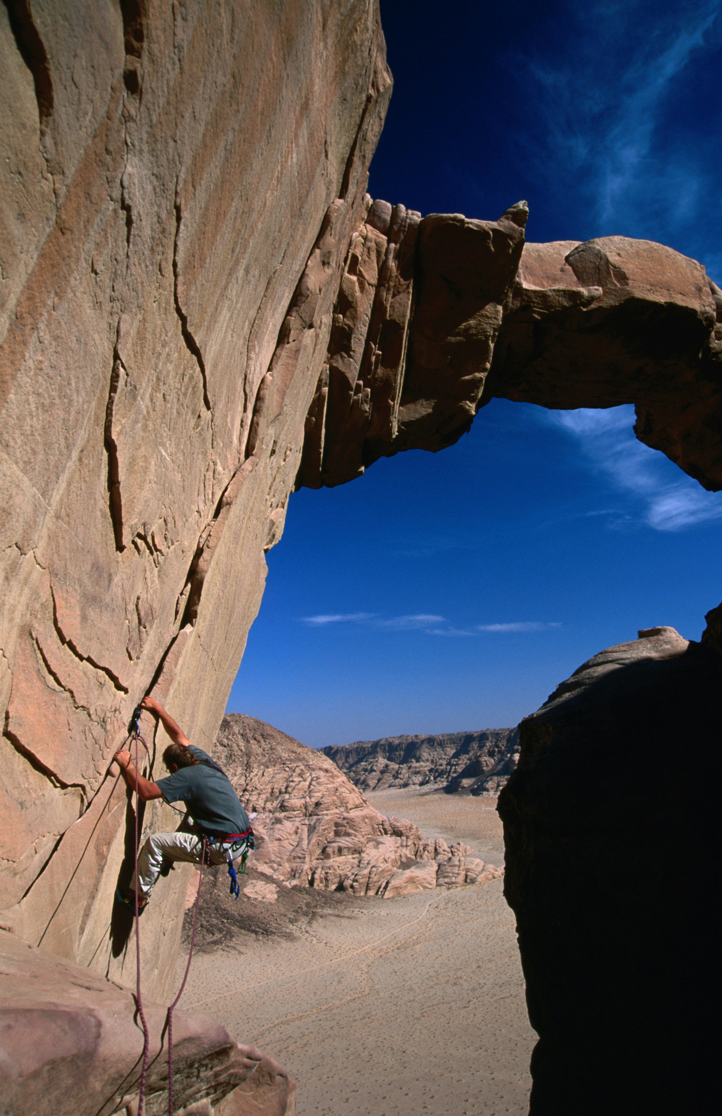 A rock climbing clings to a steeply sloping rock slab that leads up to a natural rock bridge that crests beneath a blue sky; in the distance, hundreds of feet below is the desert floor.