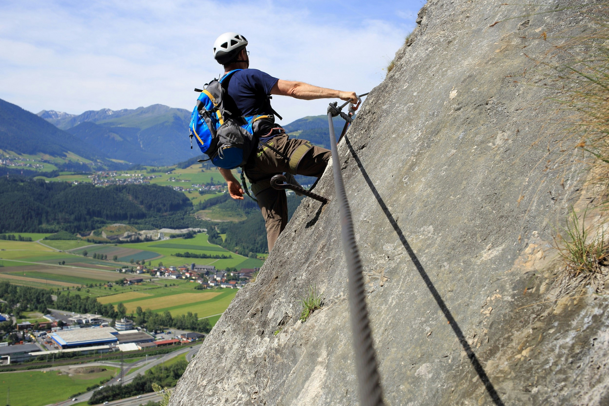 A mountaineer leans off a 45 degree rock face, with one hand extended to a steel via ferrata cable that is running horizontally along the rock; far below are green alpine valleys covered in forest and famers' fields.