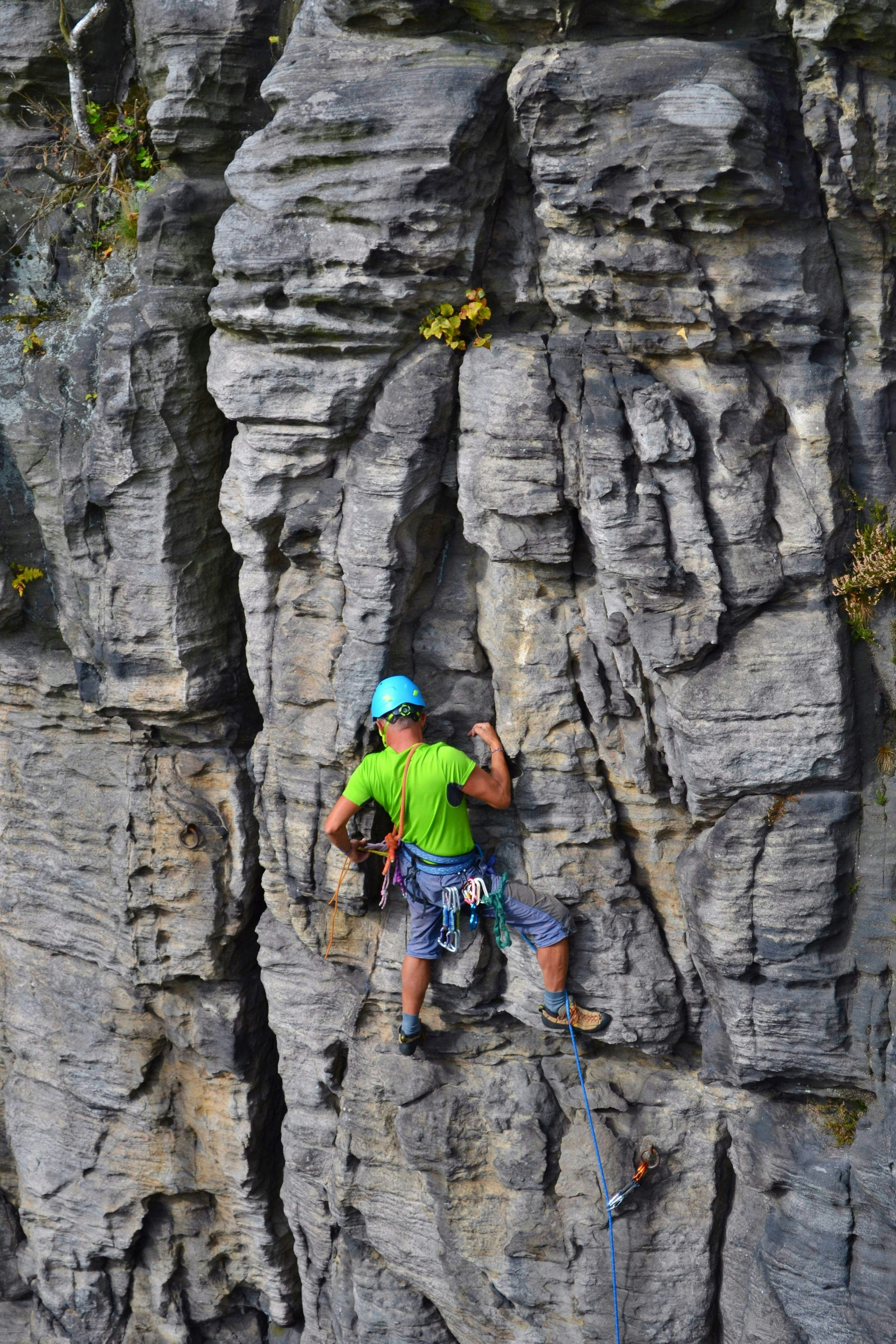 A climber in a bright green t-shirt and sky-blue helmet clings to a rock face that is full of cracks and crevices. 