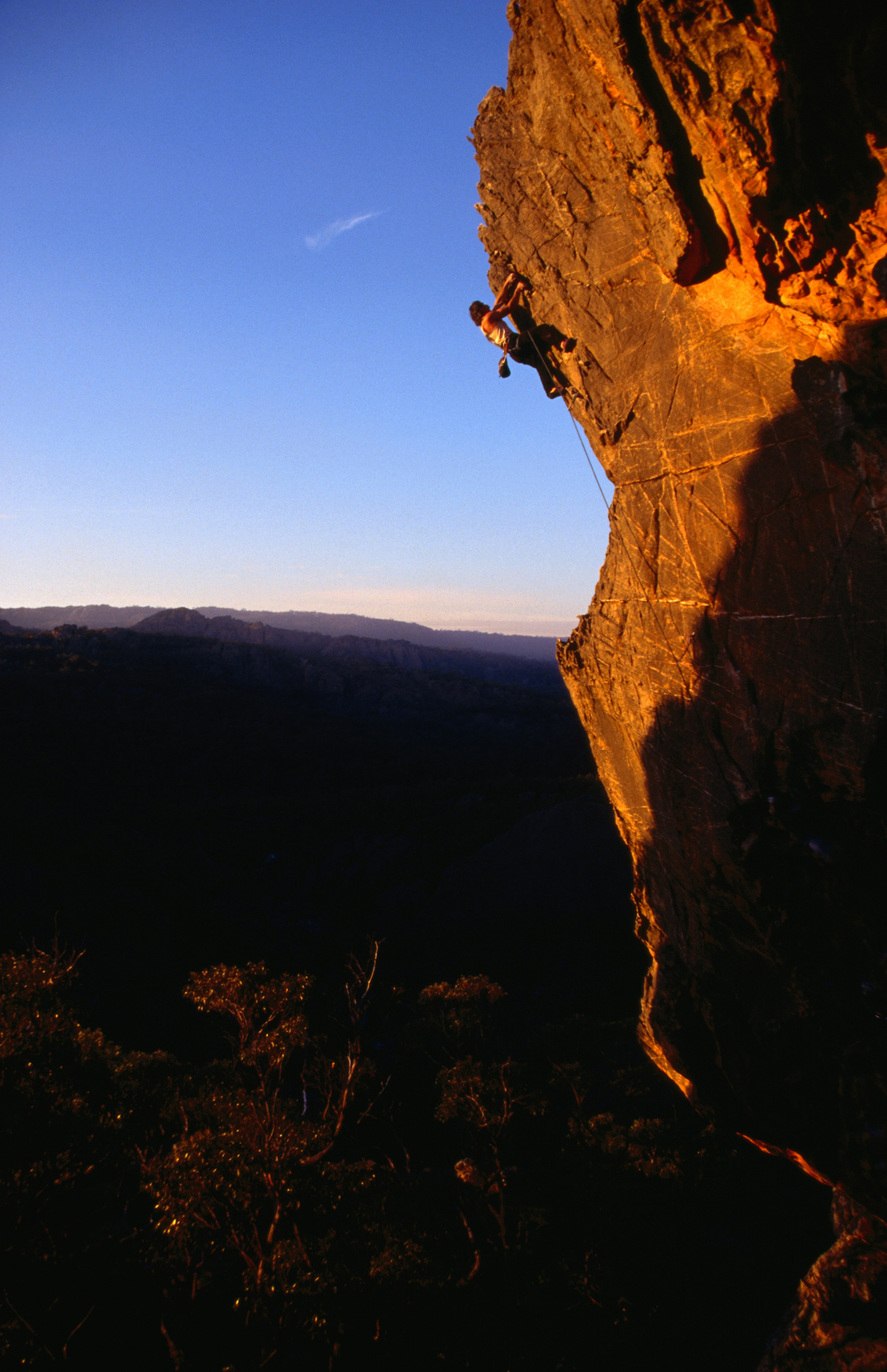 A rock climber clings to an overhanging slab of cliff face that is lit in the golden light at sunset; below him is shaded rock and behind him a sheer drop into forests far below. Above is a blue sky.