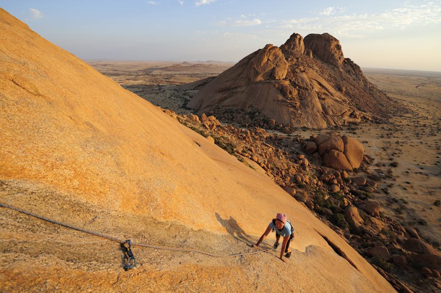 A woman climbs up a steeply sloping rock face on all fours; she is clipped into a safety rope. Far below is the desert landscape of Damaraland, with more granite monoliths rising in the distance.