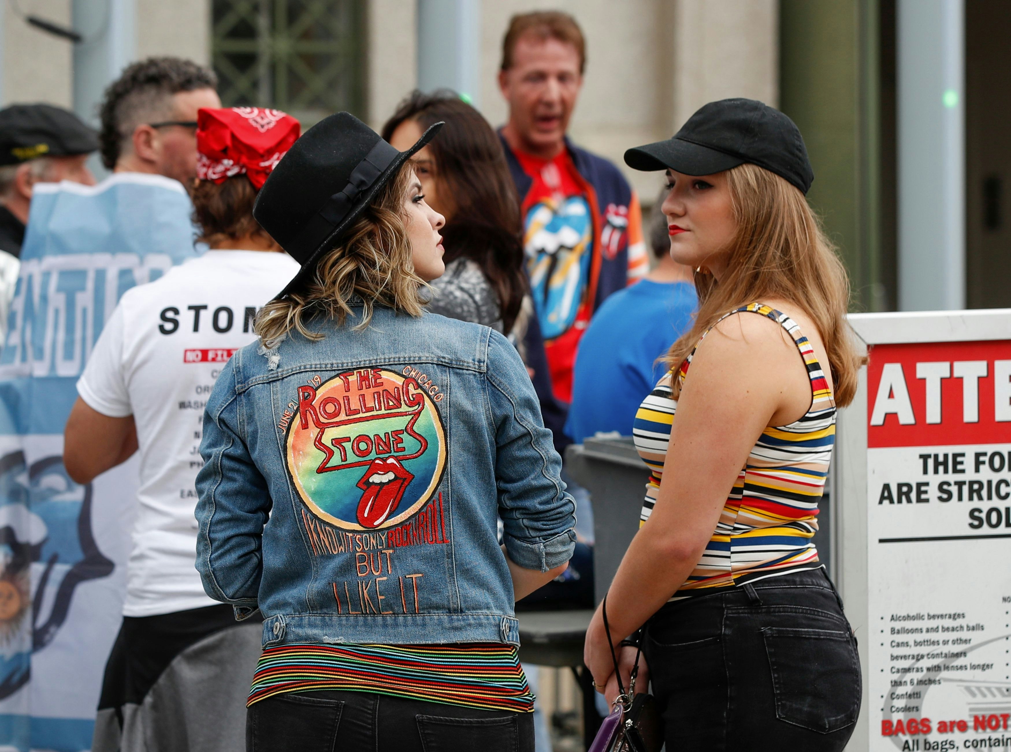 A pair of women in black jeans and black hats stand outside Soldier Field in Chicago ahead of a Rolling Stones concert. One has her back to the camera and is wearing a denim jacket embroidered with the Rolling Stones tongue logo and the lyric "I know it's only Rock N Roll But I Like It"