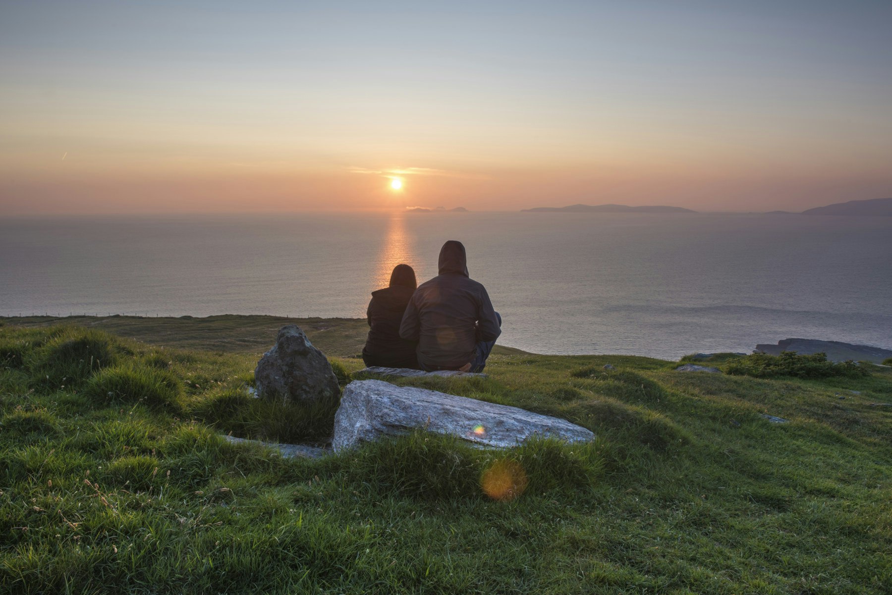 A couple sitting on a boulder on a hill watching the setting sun over the ocean