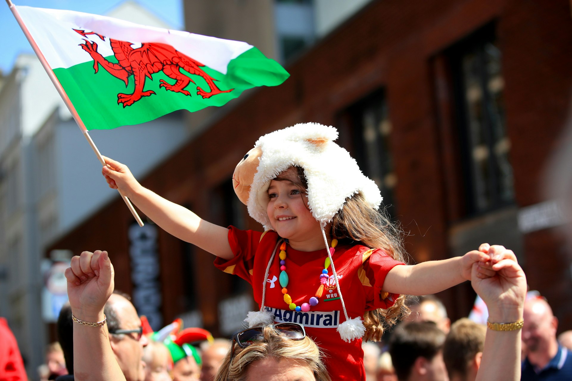 A young rugby fan, sporting a red Wales jersey and waving a Welsh flag, is carried on an adult's shoulders on a sunny day in Cardiff