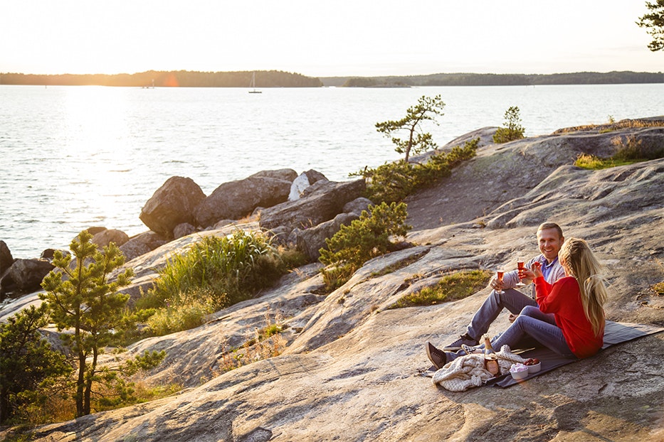 A couple enjoy a picnic on the the island of Ruissalo. They're sitting on a blanket on a rocky clearing, raising their glasses for a toast.