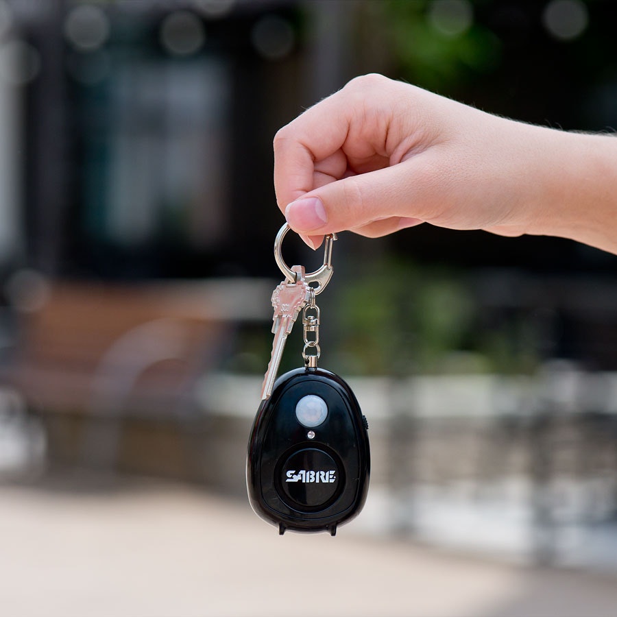 A person is holding up a SABRE personal alarm; it's black and affixed to a keyring, which also has a key on it.