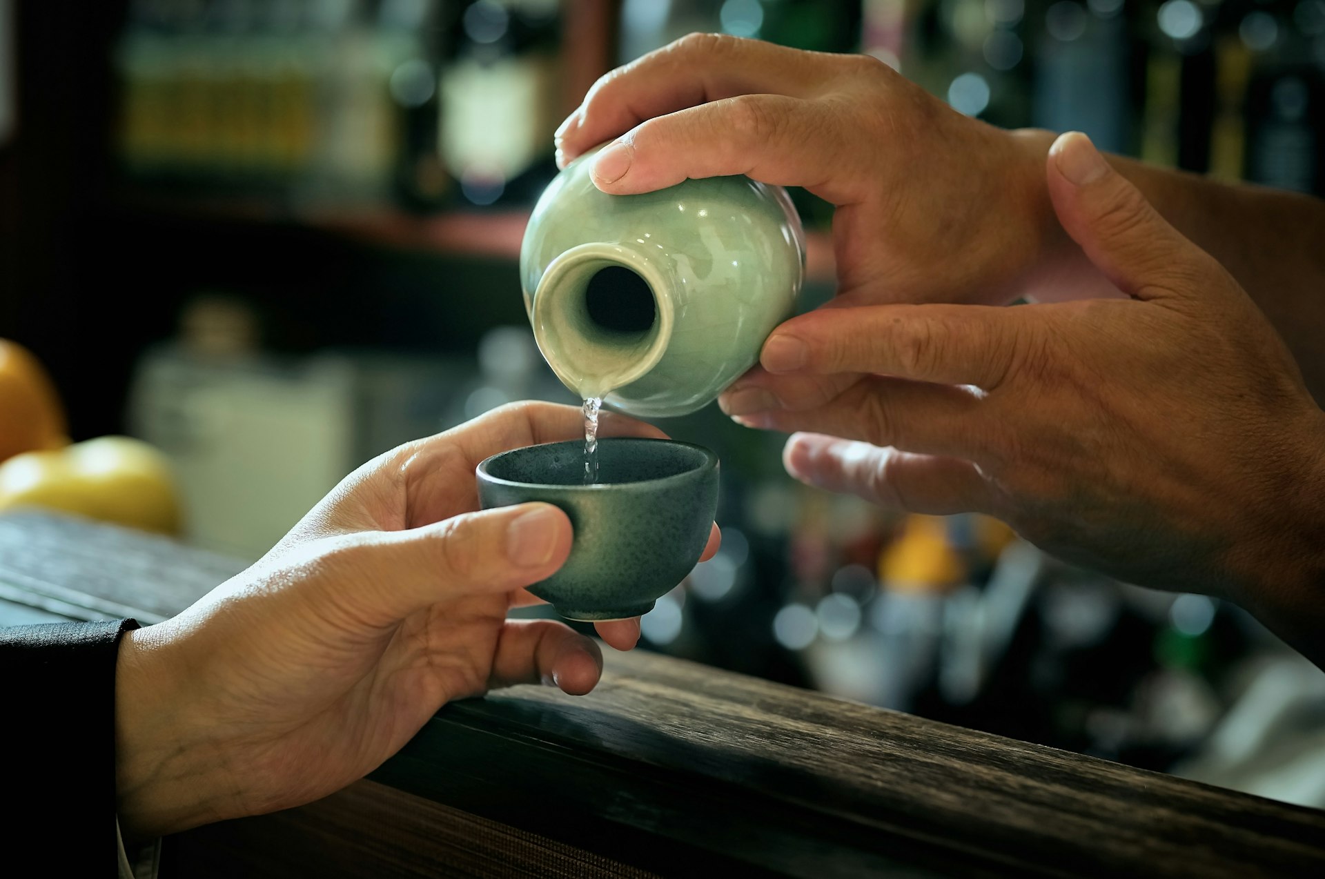 A close-up shot of a bartender pouring sake from a jug into a small cup held out by a patron.