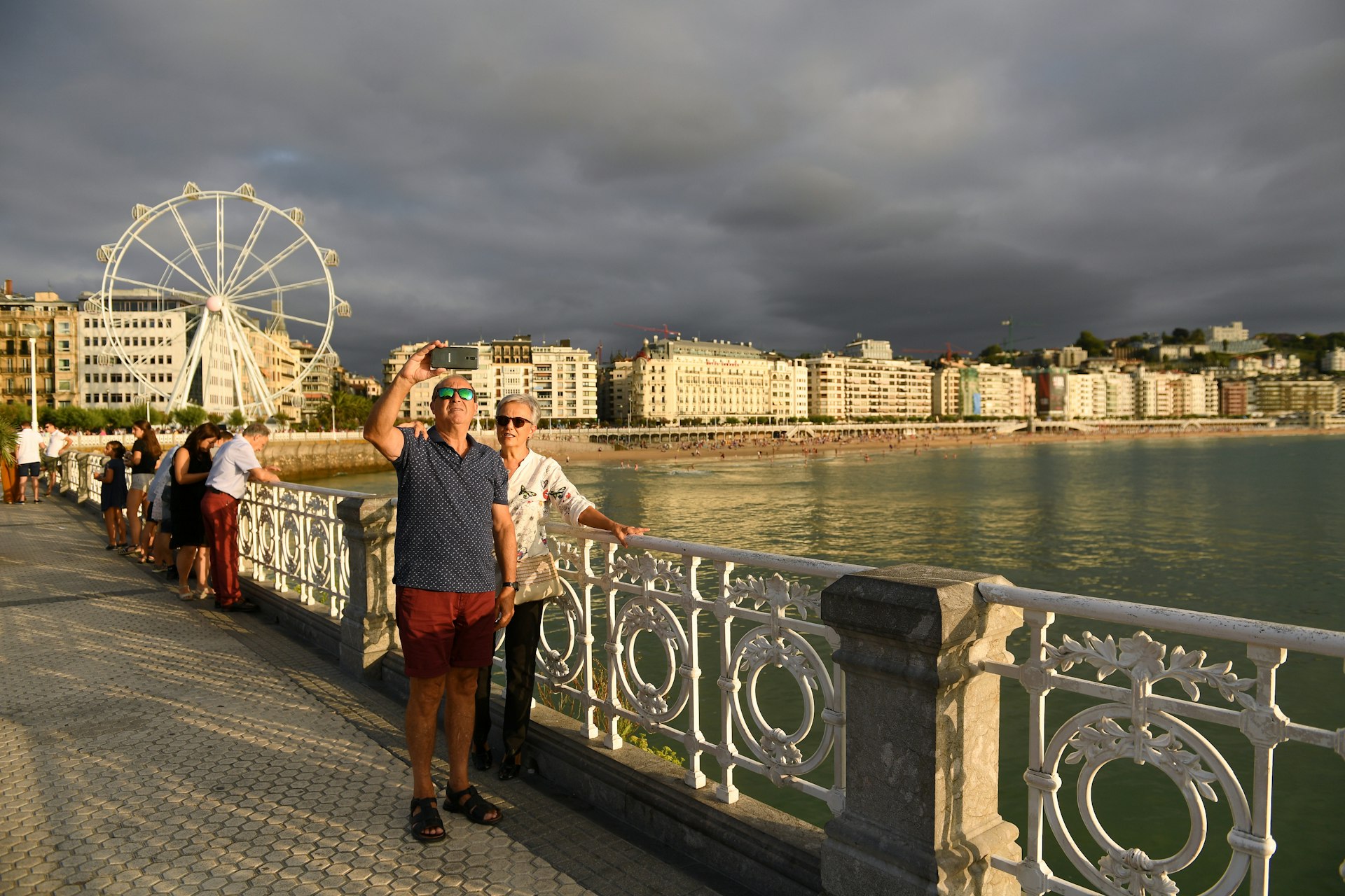 A middle aged tourist couple, the man in a blue button-down and red shorts. the woman in a white blouse and dark slacks, stand by the white metal railing overlooking the ocean on Playa de la Concha. In the background is a long strip of white hotels illuminated by the afternoon sun cutting through dark grey clouds. A ferris wheel appears on the left side of the frame. 