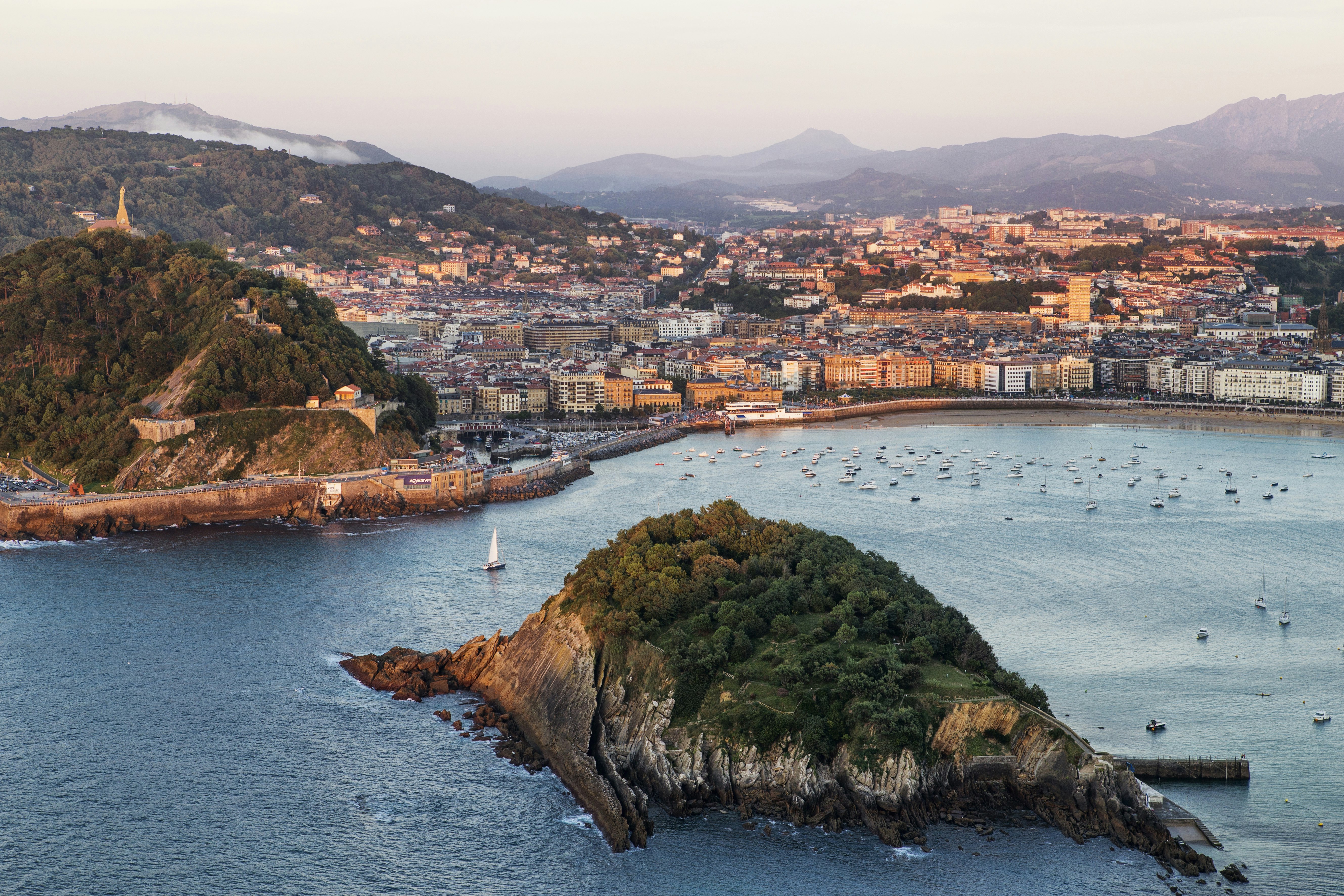 A skyline shot of San Sebastian's coastline and beachfront, with a large rocky island front and center. Buildings with a warm afternoon pink sunset glow spill across the relatively flat beach line surrounded by blue misty hills. Small boats and one large sail boat sit on the water, which is a stonewashed denim blue.