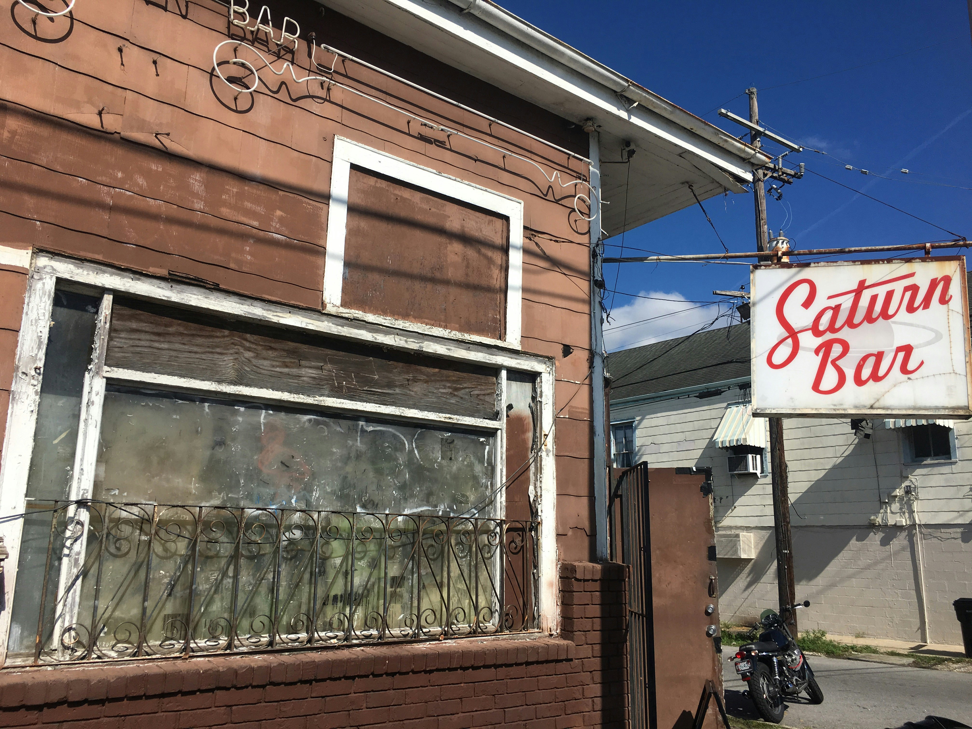 En rostig stolpe håller upp en skylt där det står "Saturnus Bar".  Barens nedgångna exteriör har bräda fönster och avskalande vita bräder.