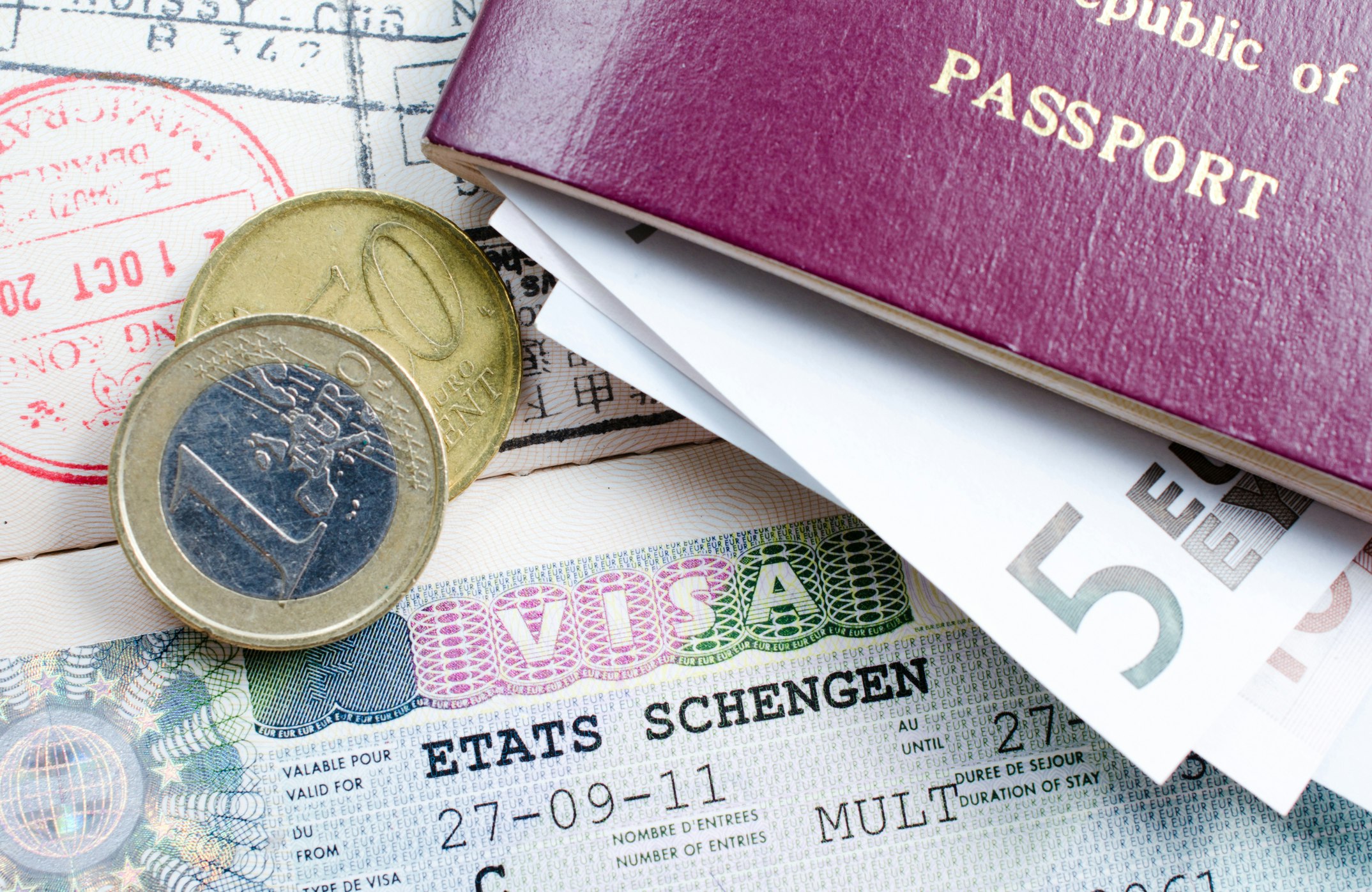 A pair of Euro coins sits on a Etats Schengen visa along with a maroon passport stuffed with paper Euro bills