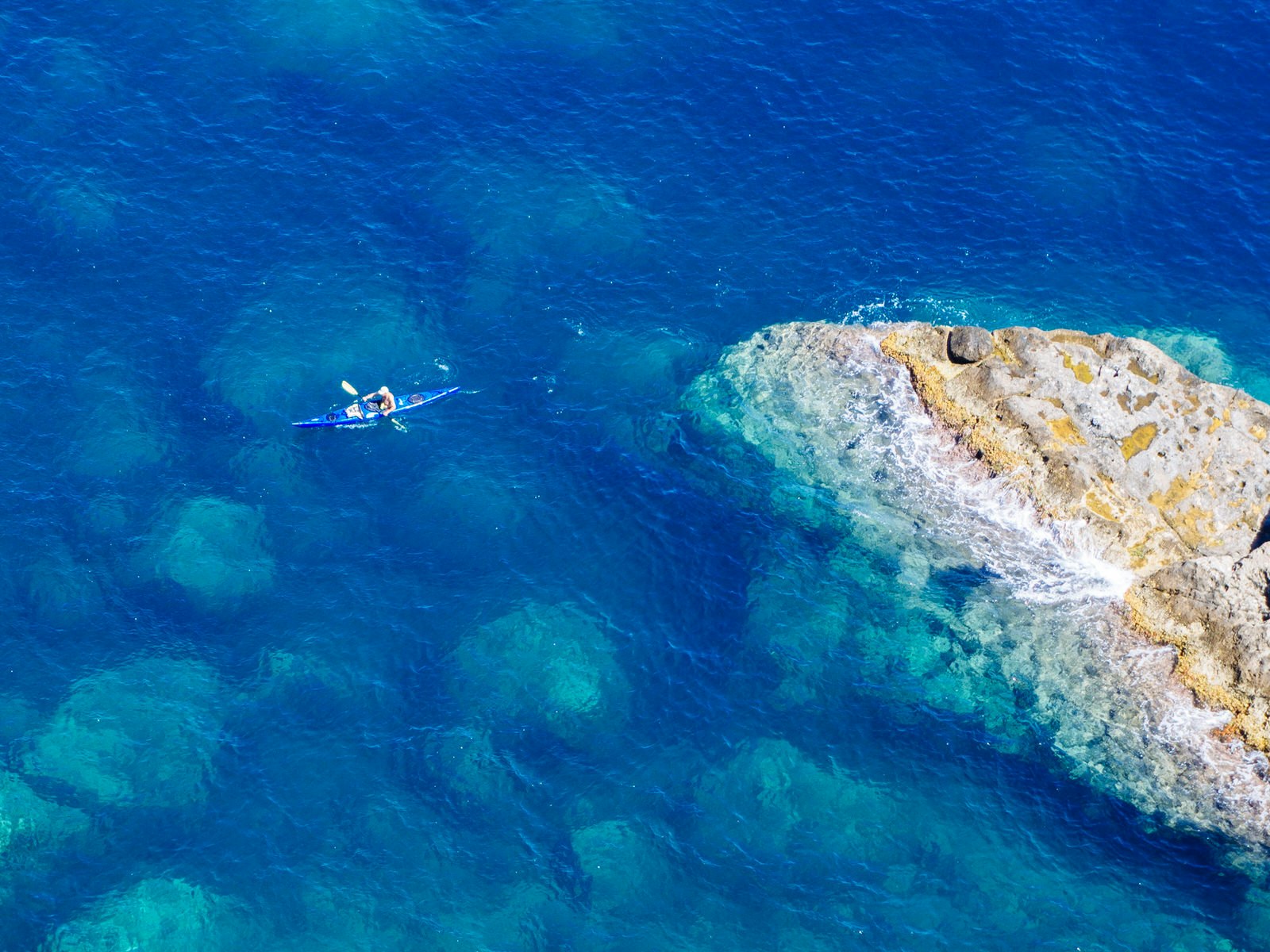 A solo kayaker surrounded by deep blue sea and a single rocky outcrop; as viewed from above.