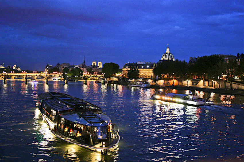 Nighttime river cruise in Paris A boat makes its way along a wide river at night. The bridge across the river and the buildings lining the shore are lit up.