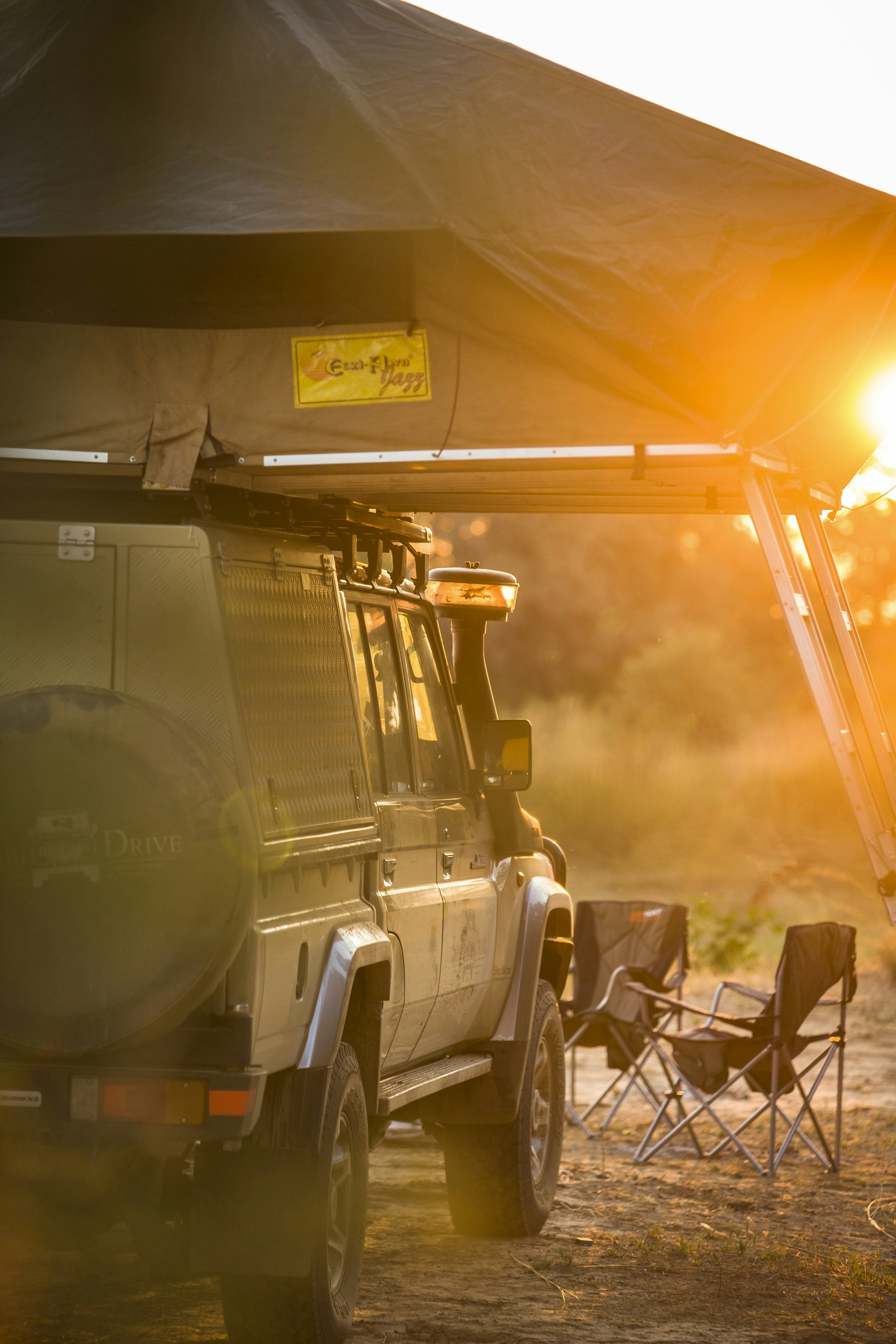 A self-drive 4WD vehicle is parked in a campsite, with folding chairs in front and a tent on the roof. The sun is setting and the light is golden.