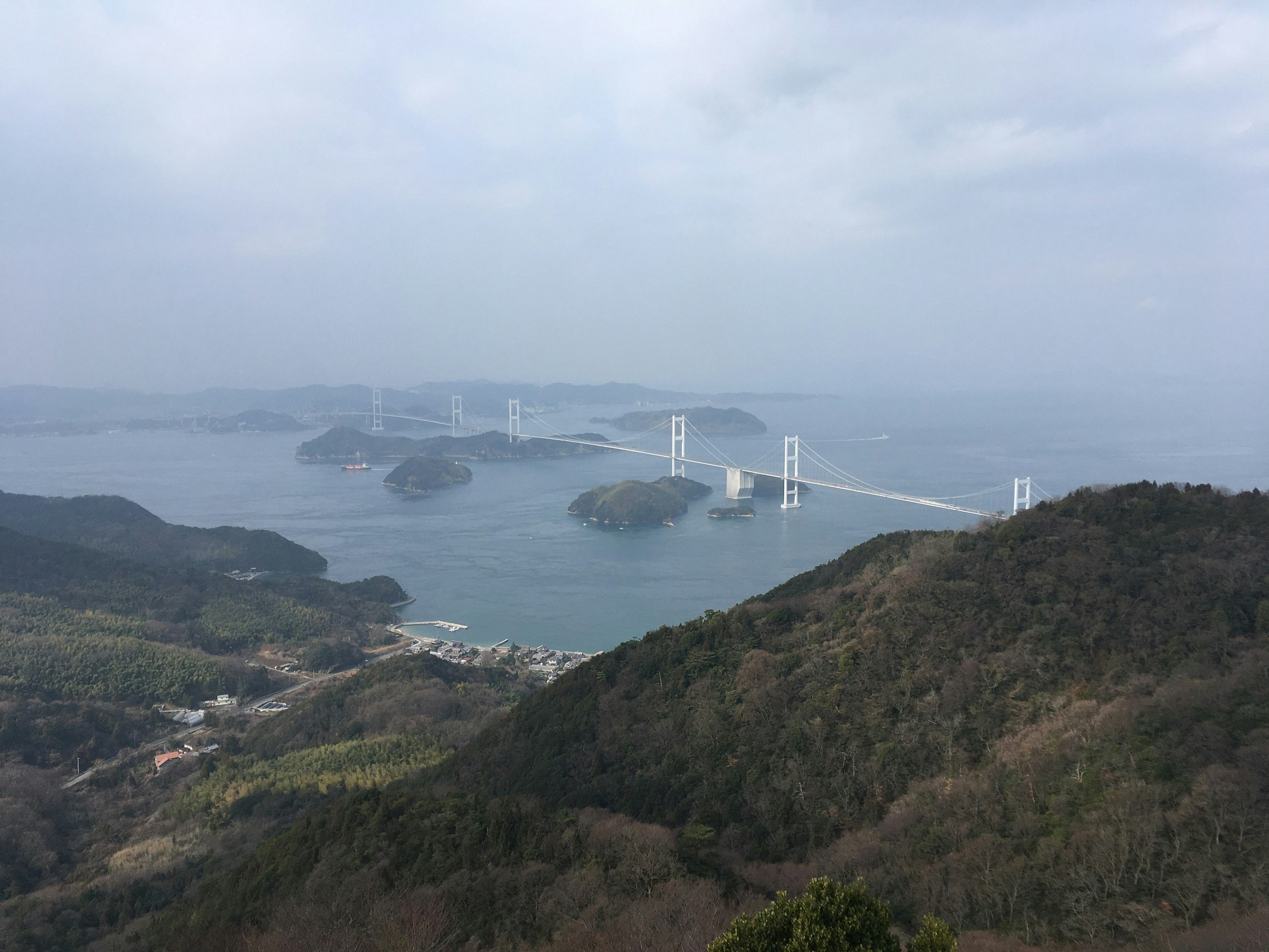 A view down hillsides to a village and across a sea to a large white suspension bridge stretching into the distance.