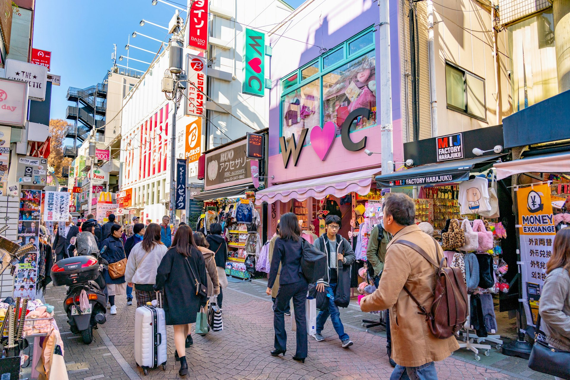 A side-on view of Takeshita-dori: a pedestrian shopping street lined with fashion boutiques, cafes and restaurants. Many people walk along through the streets, browsing the shops as they go, while above their heads, signs for various businesses protrude from the walls.