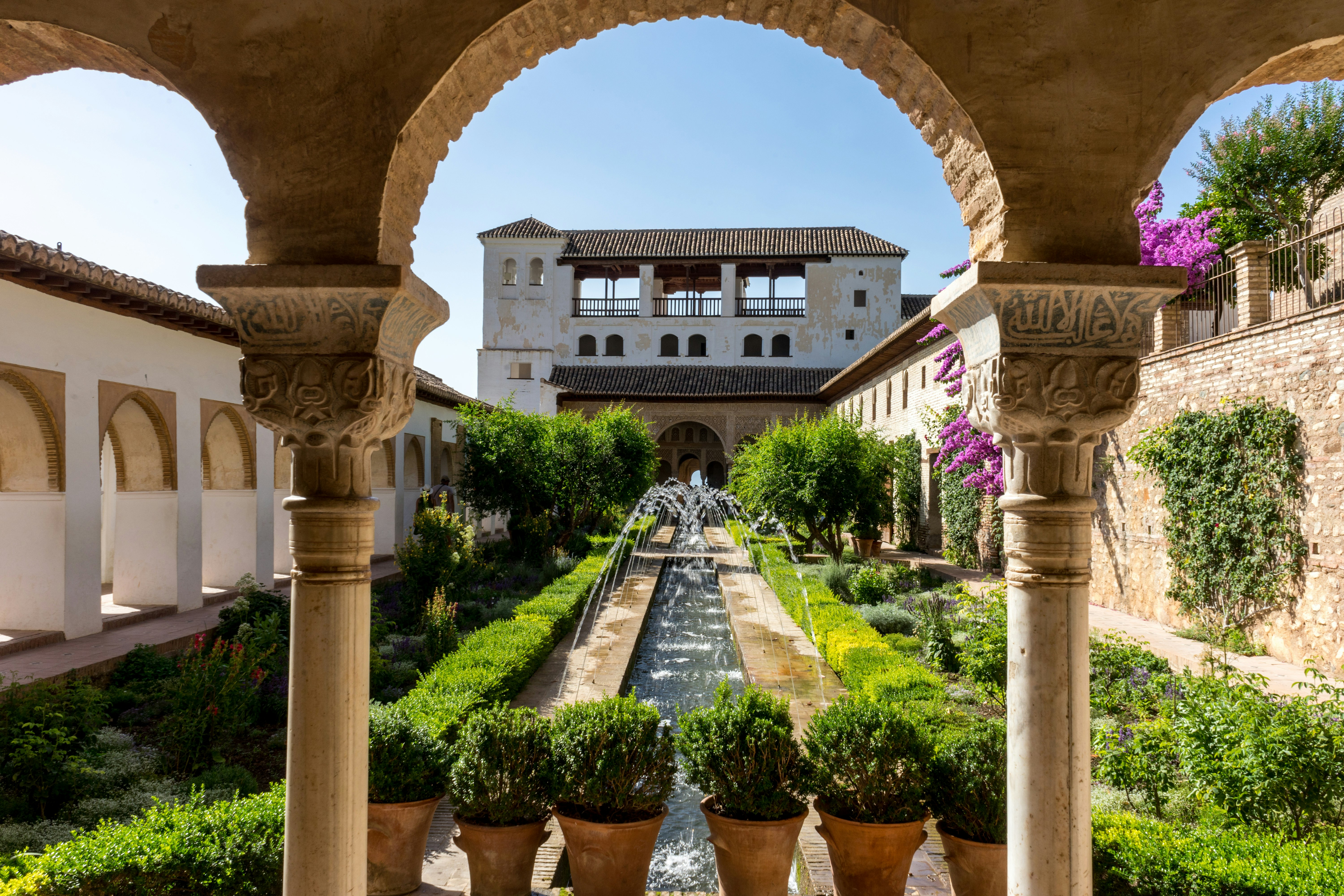 Looking between the columns of an archway in a palace, towards a walled garden. There are lush green plants and a long, narrow, rectangular pond with fountains arcing over it.