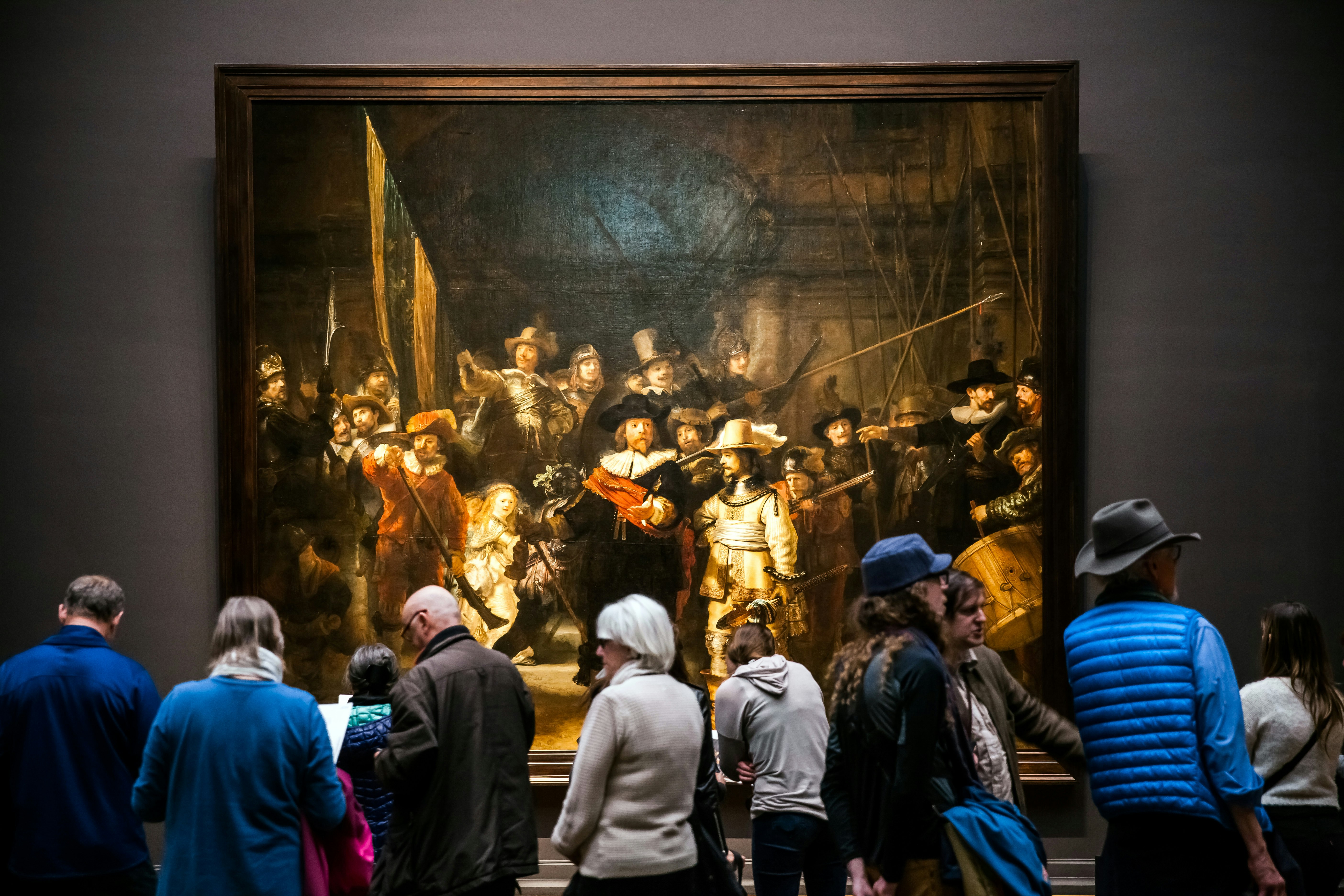 En grupp människor står framför 'The Night Watch', en stor, berömd målning av Rembrandt, hängande i en storslagen ram.