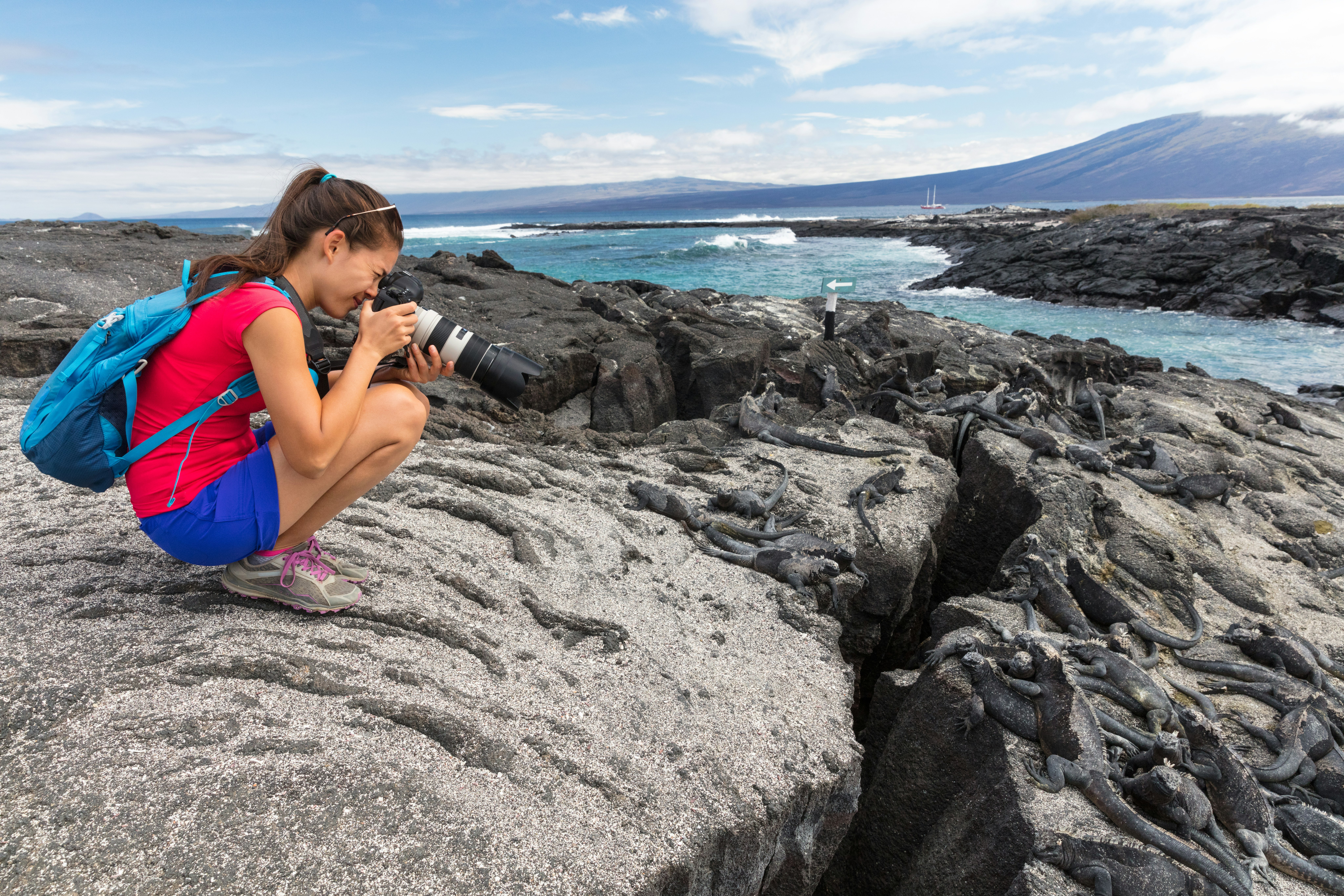 A young brunette woman with a ponytail, red t-shirt, blue shorts, light blue backpack, and grey tennis shows squats on a rock bluff taking a picture of the surrounding ocean scenery using a DSLR camera with a long telephoto lens 