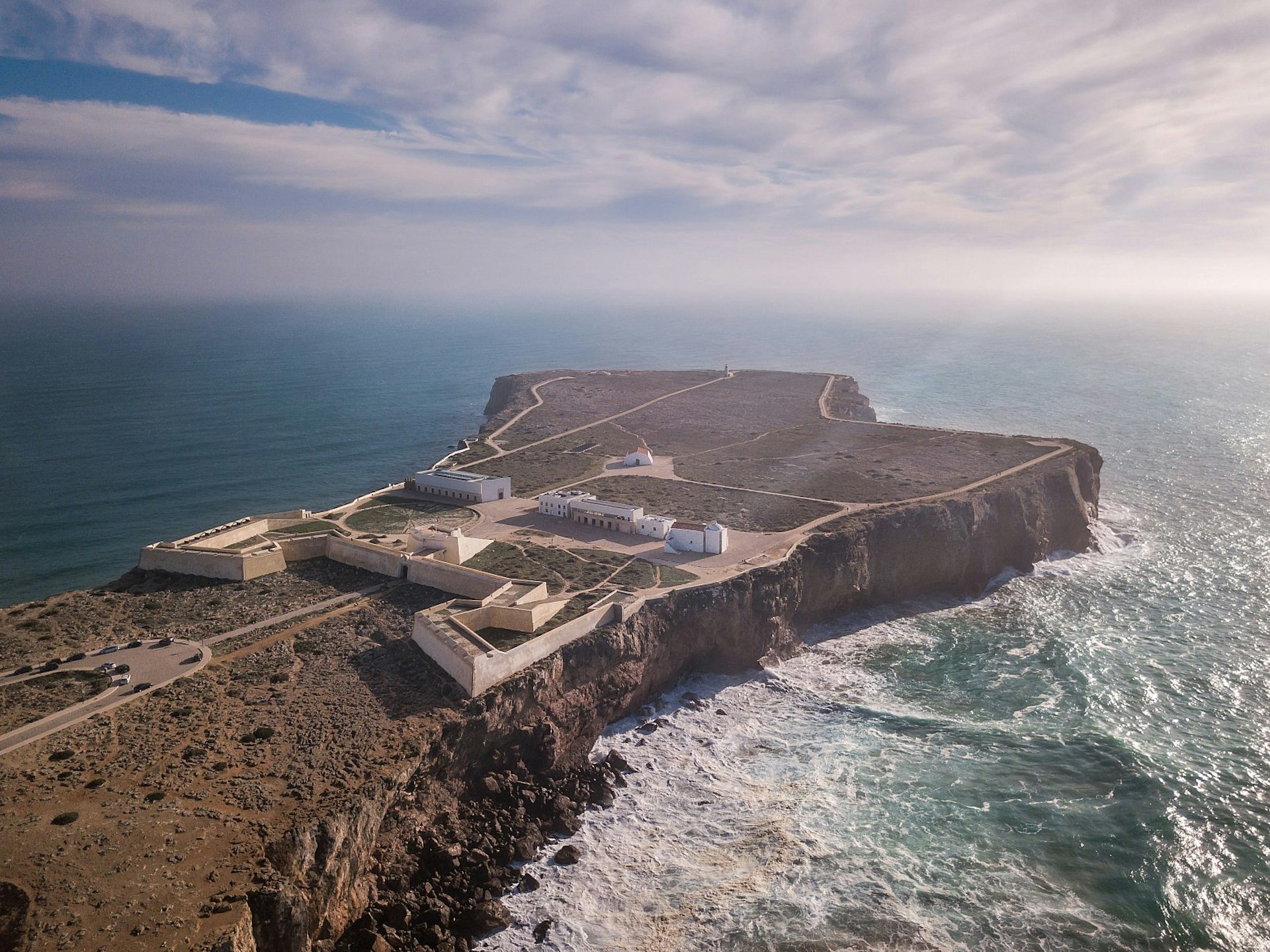 An aerial view over a fortress in a dramatic location at the end of a promontory; heavy waves crash against the cliffs below. 