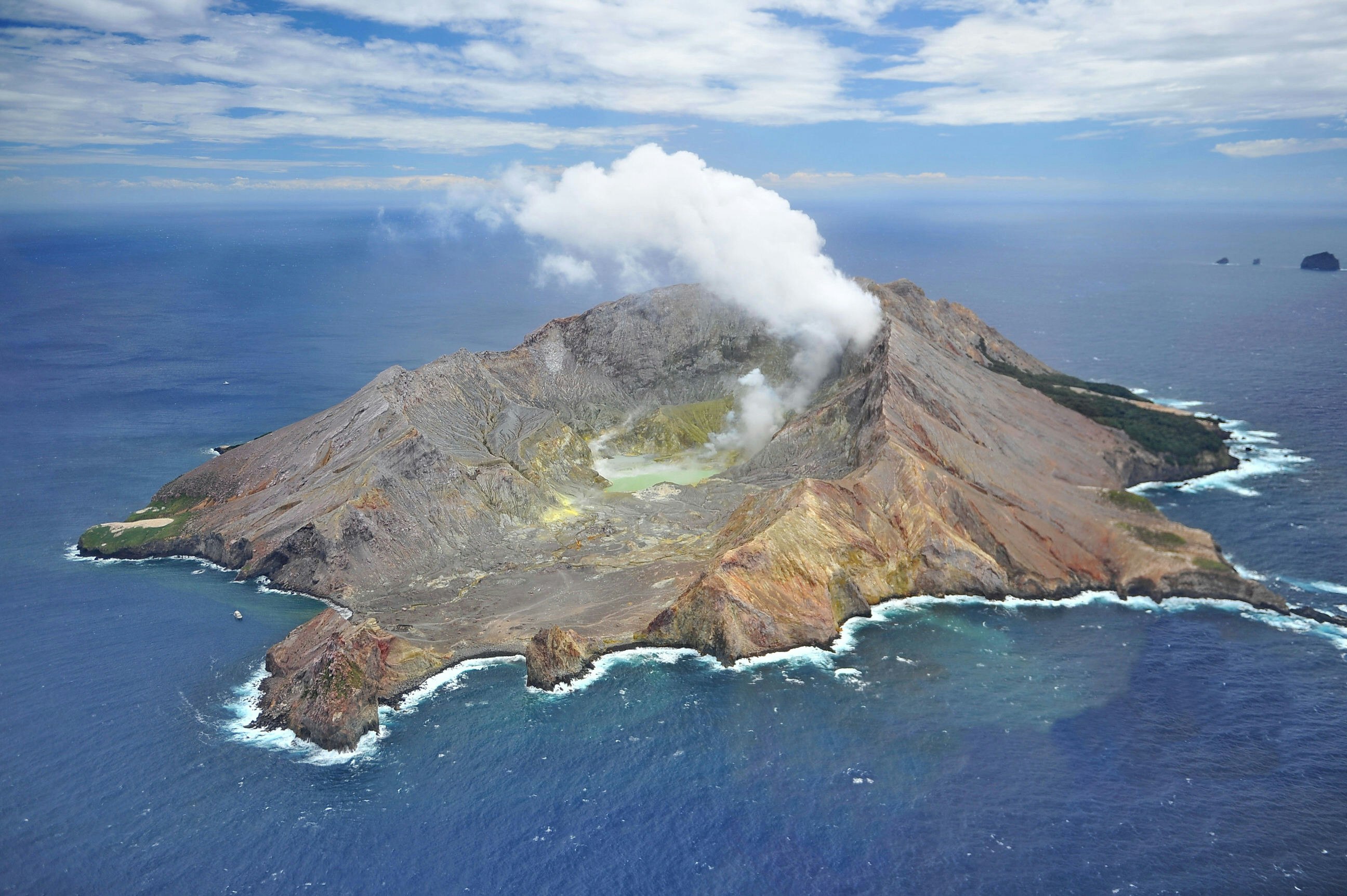An aerial view of a craggy, volcanic island. There is steam rising from the top, and a green crater lake in the middle. The island is surrounded by dark blue sea.