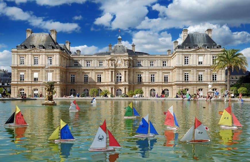 Colourful wooden toy boats sail on the still waters of the pond in Jardin du Luxembourg, Paris. There is a stately building with beige bricks in the background underneath a clear blue sky. 