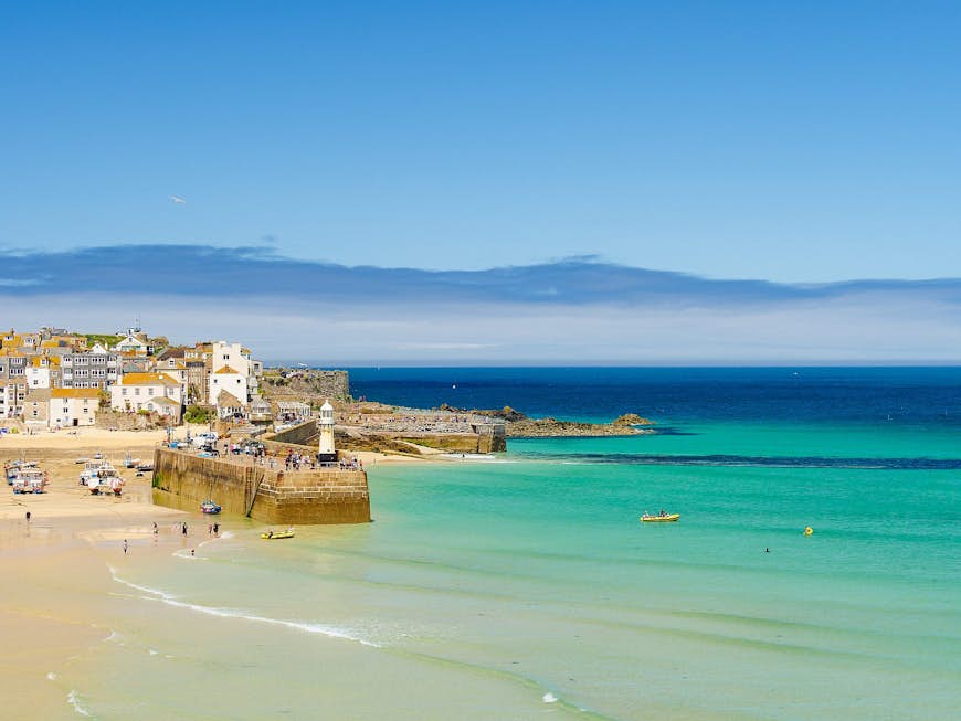 The beach and clear turquoise waters of the Atlantic at St Ives, with the white cottages of the town spilling onto a headland beyond the harbour wall.
