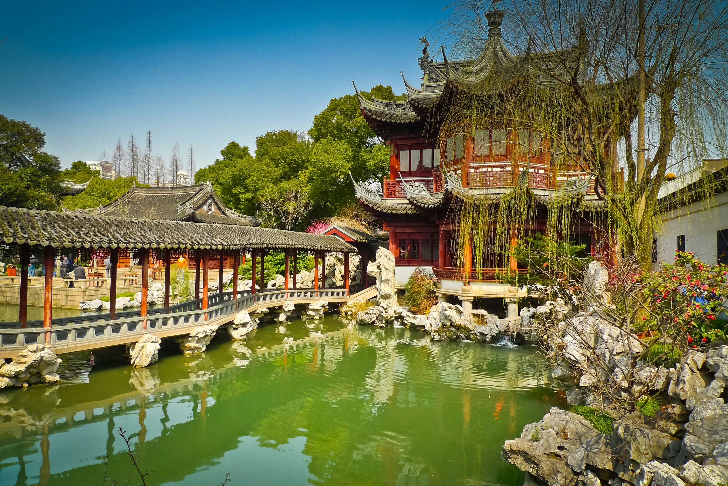 A traditionally styled bridge spans across a pond in Yuyuan Gardens in Shanghai. On the far edge of the pond stands a Chinese temple, surrounded by shrubbery.