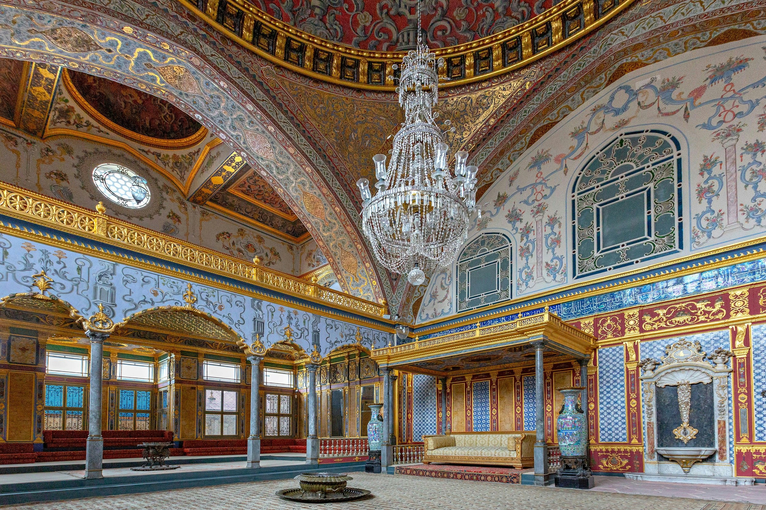 A grand room covered in ornate mosaic patterns, predominantly in gold, red and blue. A large chandelier hangs in the centre of the room