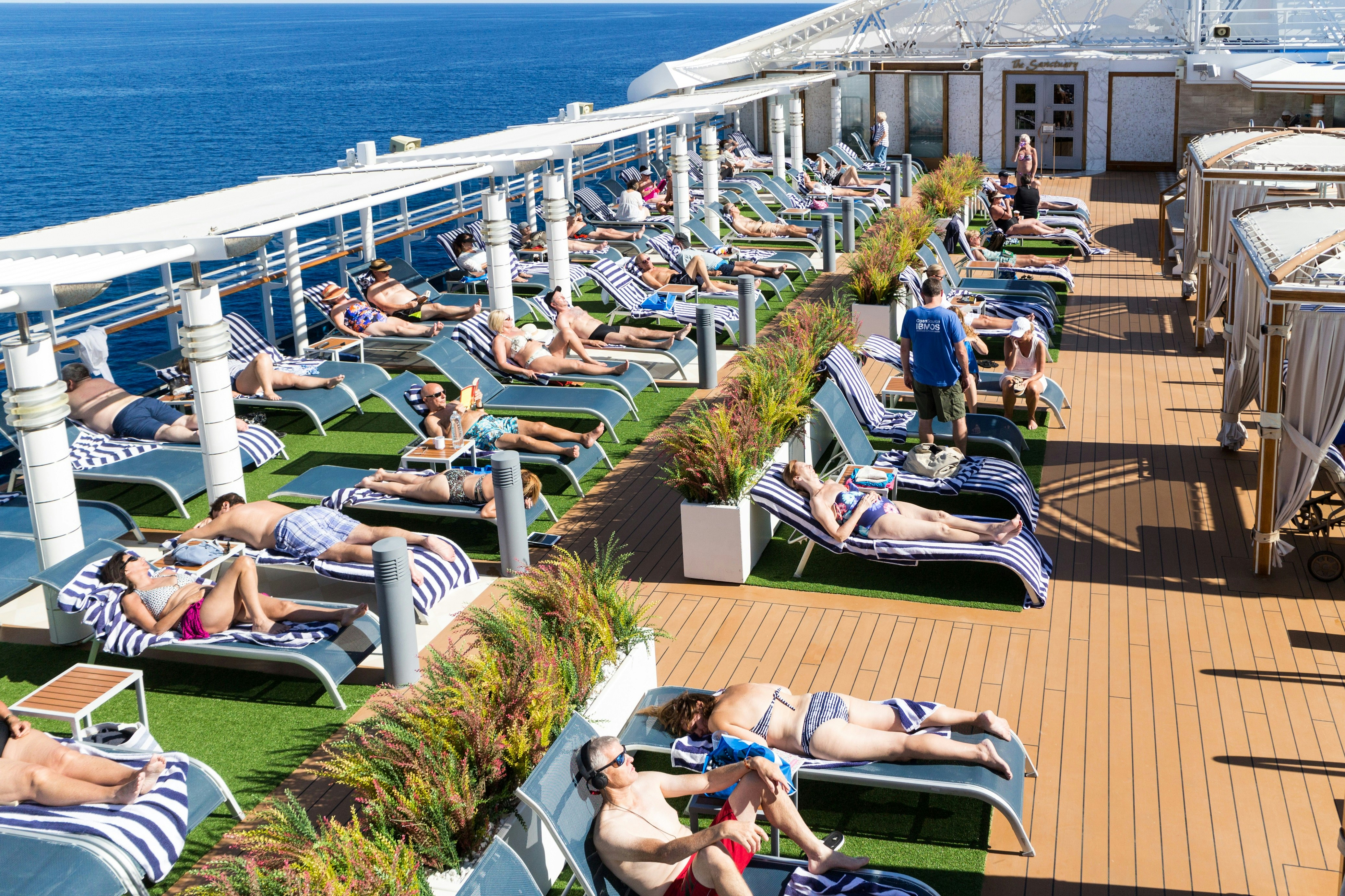 Passengers sun themselves on blue sun loungers lined up along the deck of a cruise ship. The boat's edge is visible with the blue sea beyond.