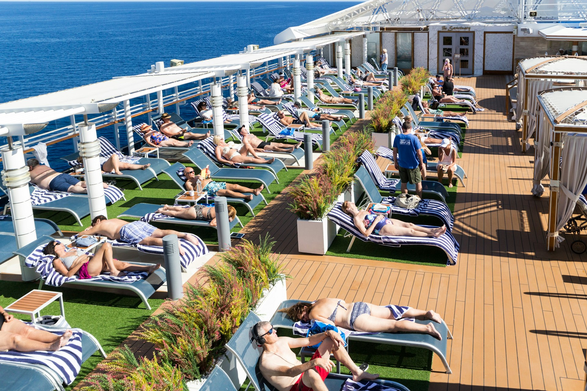 Passengers sun themselves on blue sun loungers lined up along the deck of a cruise ship. The boat's edge is visible with the blue sea beyond.