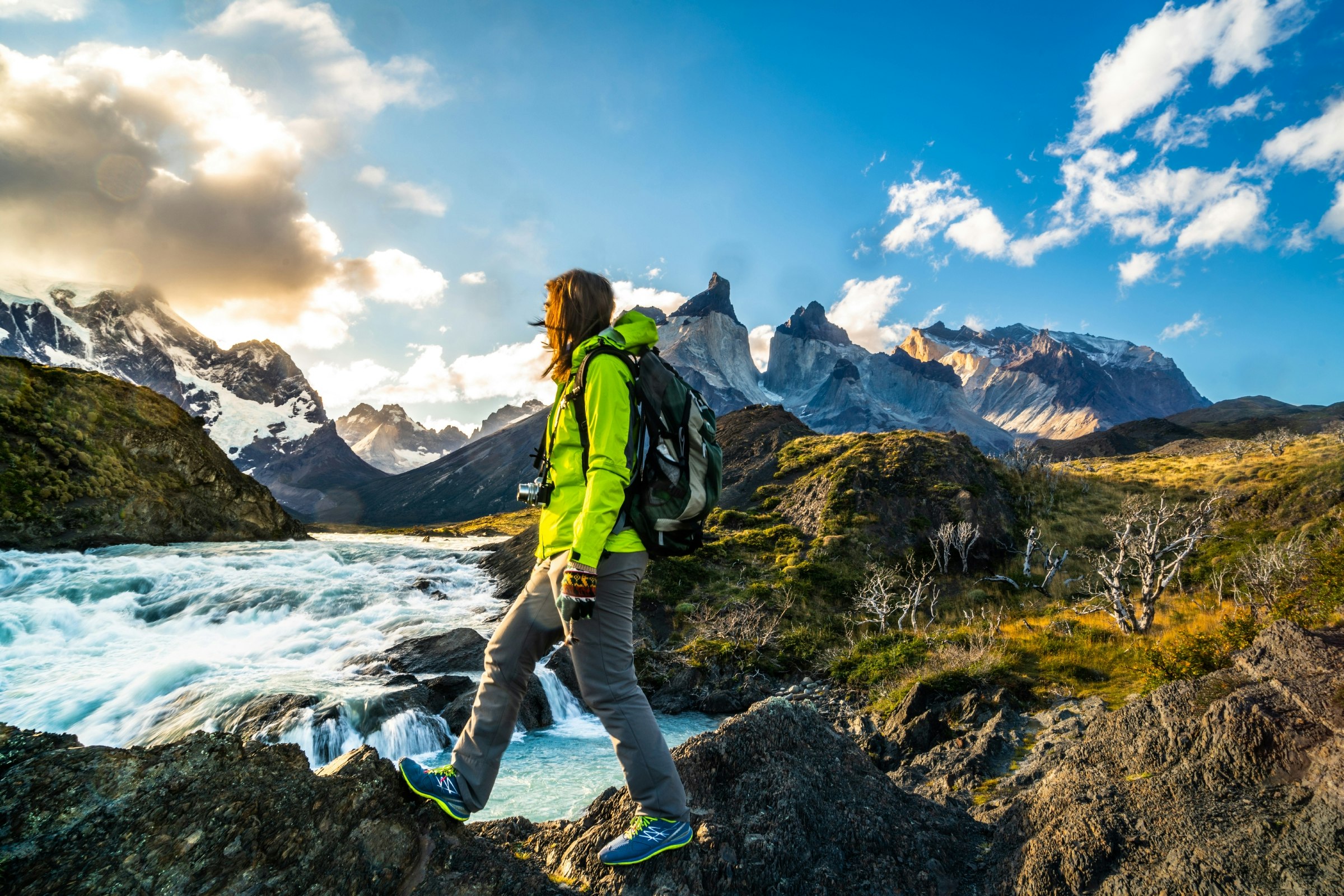 A female hiker walking on rocky ground near Salto Grande Waterfall in Torres del Paine National Park, Chile. In the background numerous snowy mountains are visible.