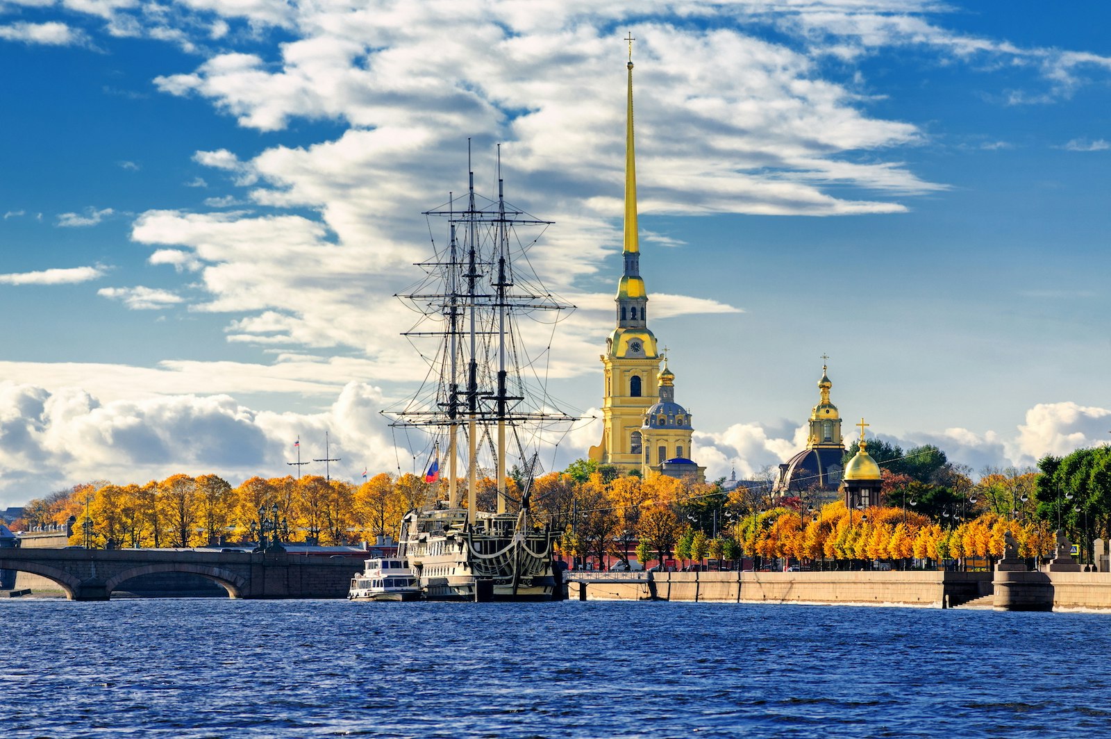 Sailing ship anchored by the Peter and Paul Fortress, with orange-leaved trees lining the coast