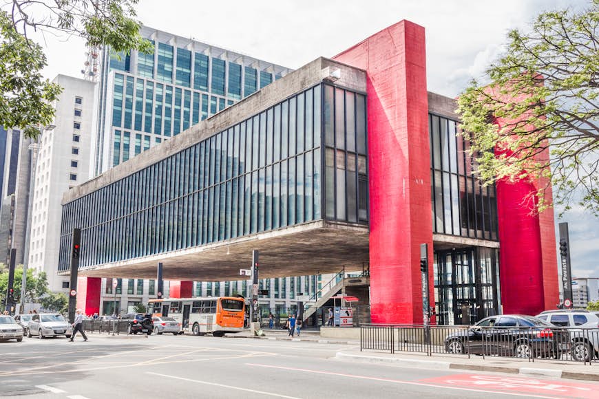 A concrete building with many windows raised from the ground on huge red blocks above a plaza busy with traffic.