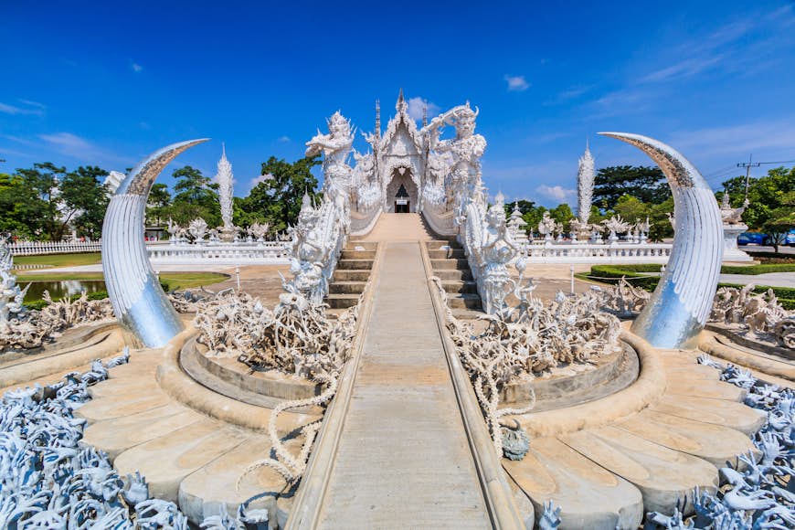 The entrance to Wat Rong Khun, an art exhibition in the style of a Buddhist temple in Chiang Rai. The narrow path to the temple entrance, which is bright white, passes over a 'lake' from which hundreds of sculptures of hands grasp upwards towards the blue cloudless sky.