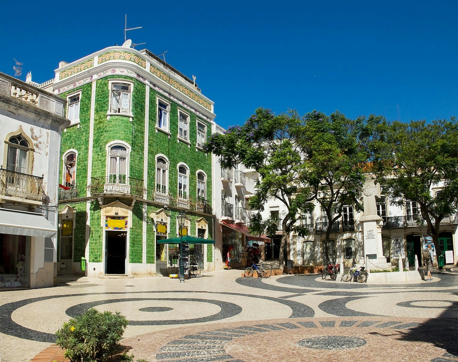 Looking over a sunlit square, paved with stones in a circular geometric pattern; the centre of attention is a handsome green tiled house, alongside white buildings and trees. 