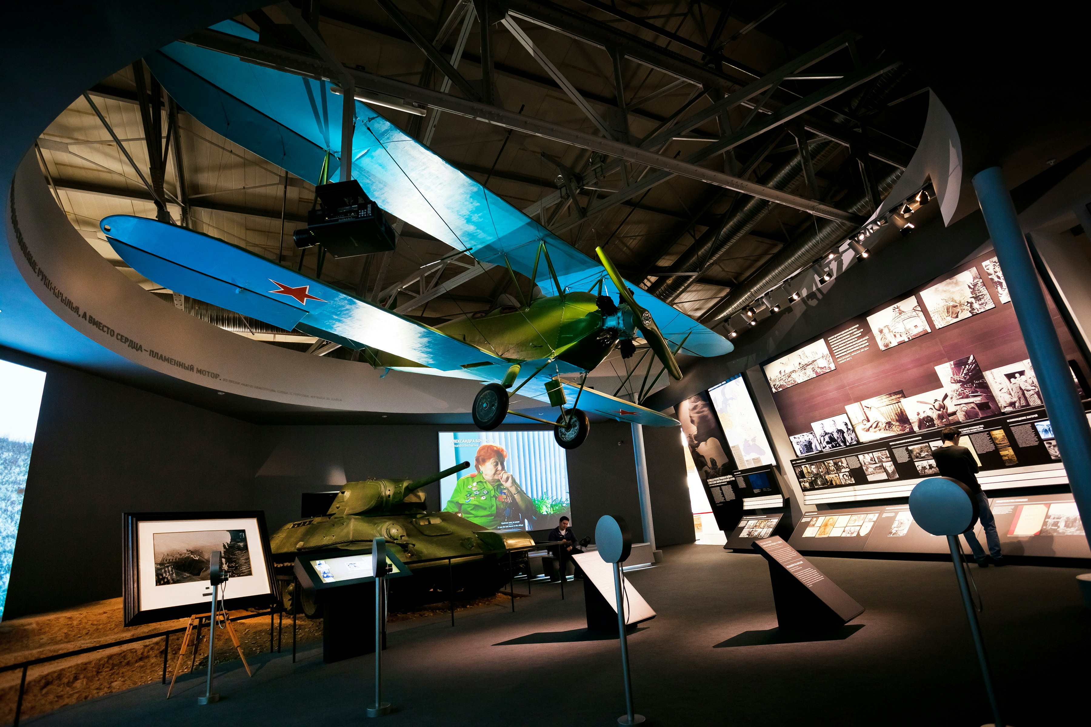 A large dark room, with many illuminated exhibits including a wall of black-and-white photographs. A tank stands against a wall and a biplane hangs from the ceiling