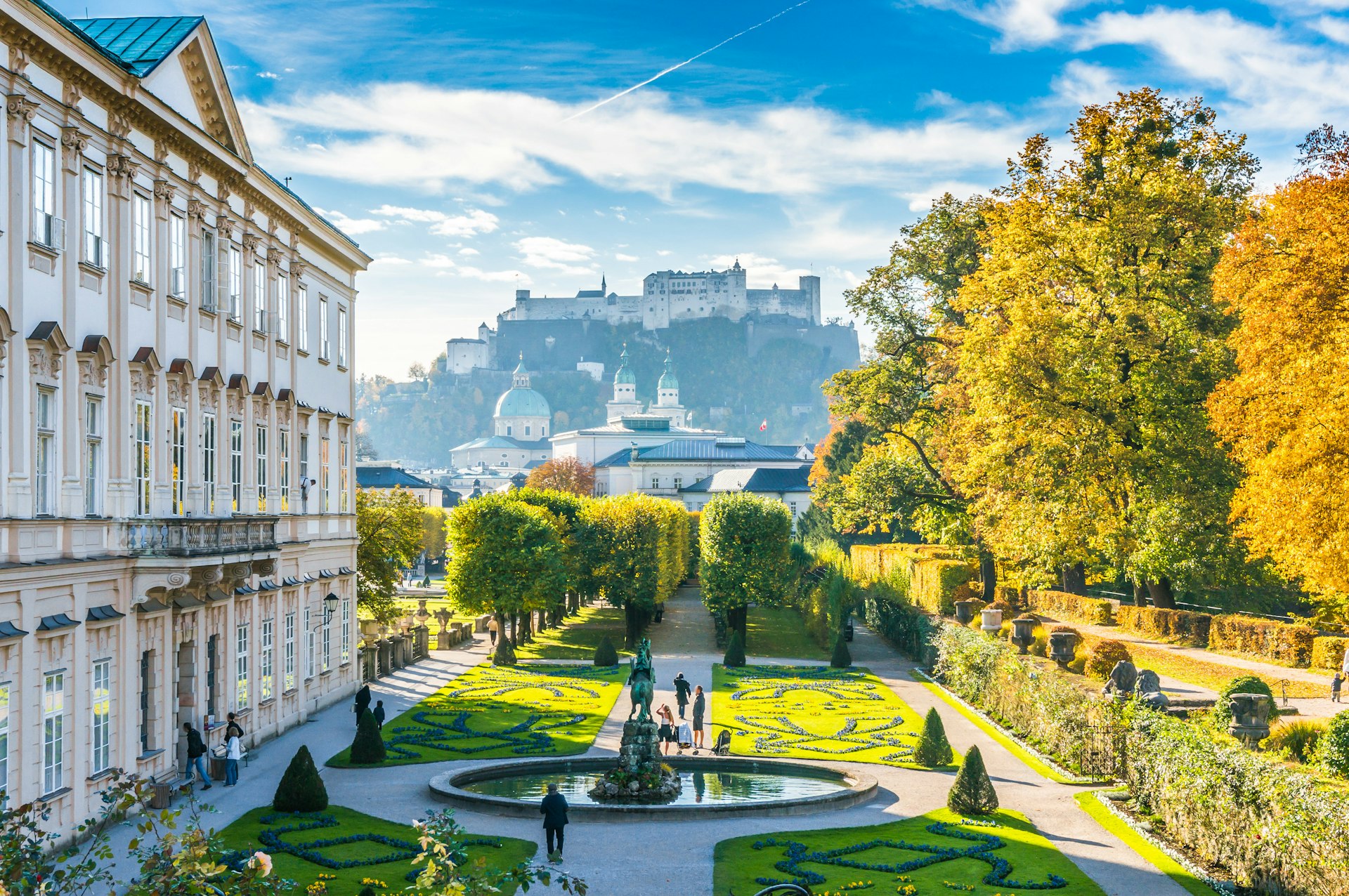 A view from Salzburg's Mirabell Gardens with the city's hilltop fortress in the background. The buildings are grand and white while the gardens are green and neatly manicured.