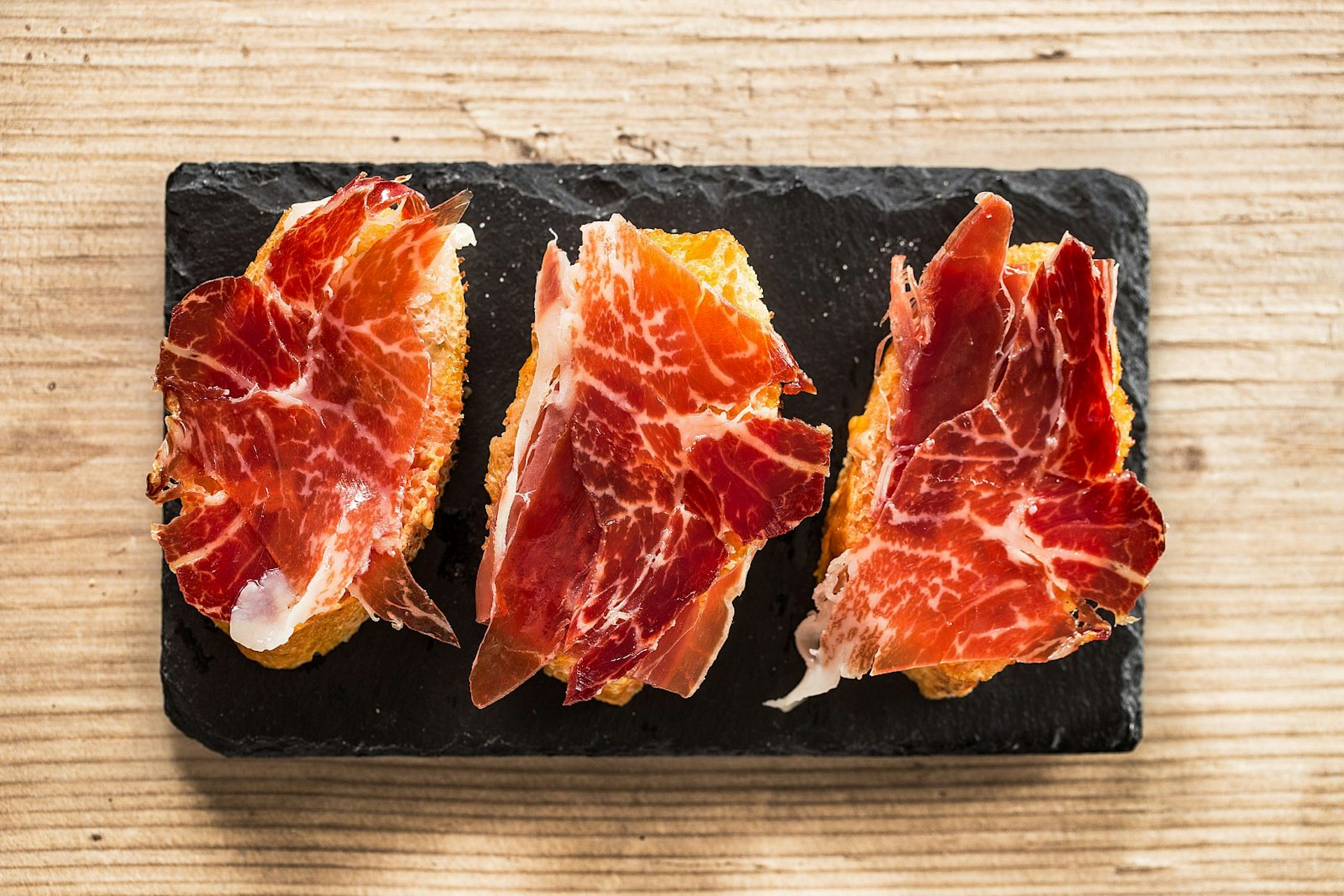 Slices of jamón ibérico on bread on a black slate on a wooden tabletop.