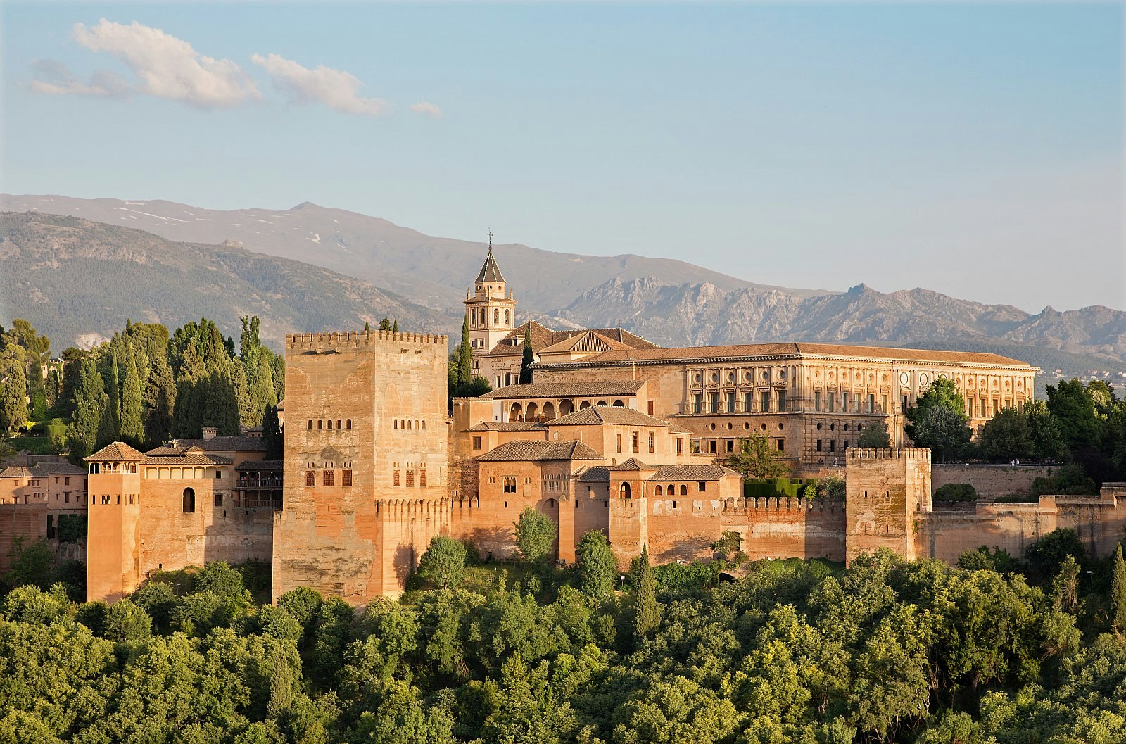 The sand-coloured Moorish palace complex of the Alhambra, with verdant trees in the foreground and stark mountains as the backdrop.