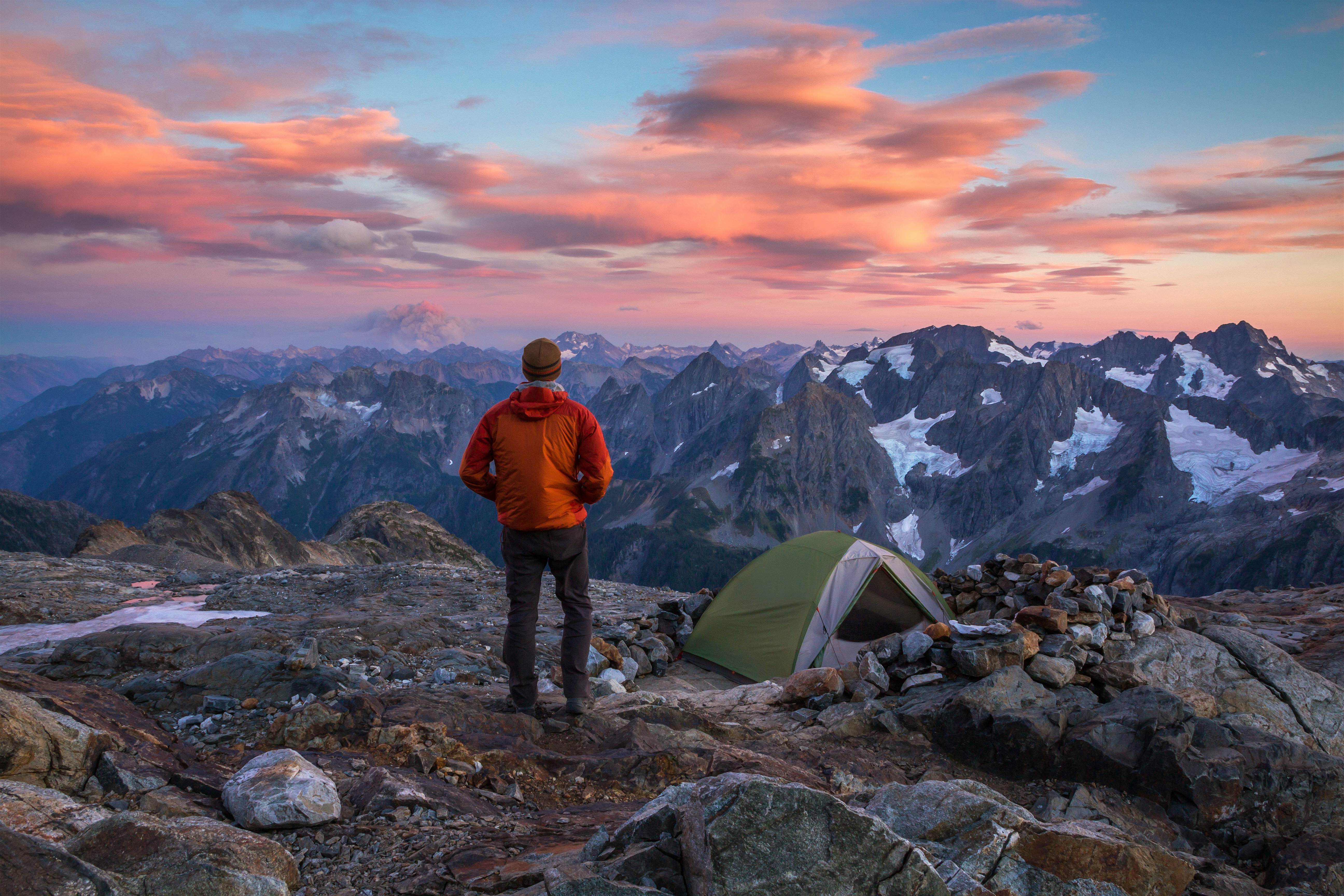 North Cascades National Park Hiker in warm clothing standing at their mountain-top camp, watching the clouds glow sunset colors above the mountains in North Cascades National Park