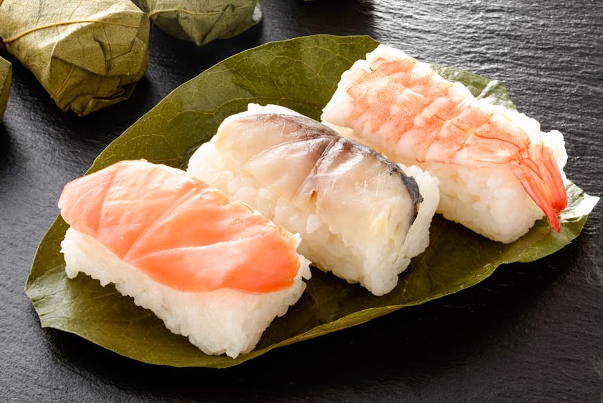 Three pieces of kakinoha sushi - one prawn, one salmon and one white fish - served on a persimmon leaf on a black slate tabletop, with more in the background wrapped in the leaves. 