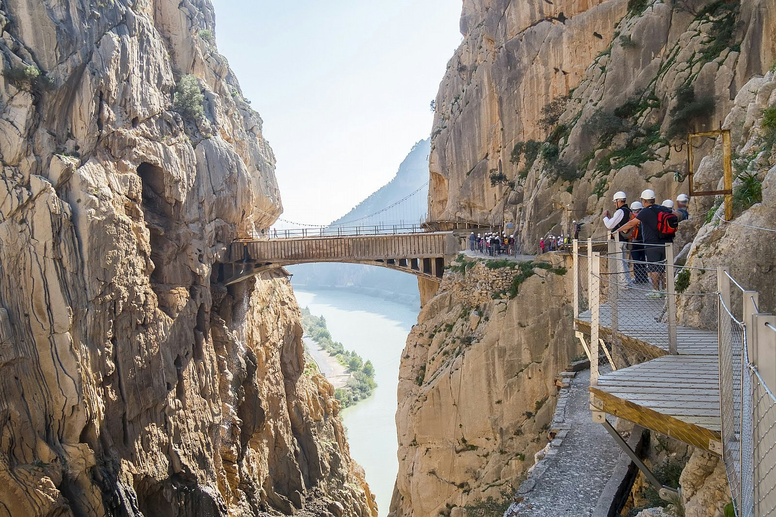People in hard hats on a boardwalk snaking around a rocky cliff; up ahead the boardwalk crosses a narrow gorge to join the cliff on the opposite side, with a river far below.