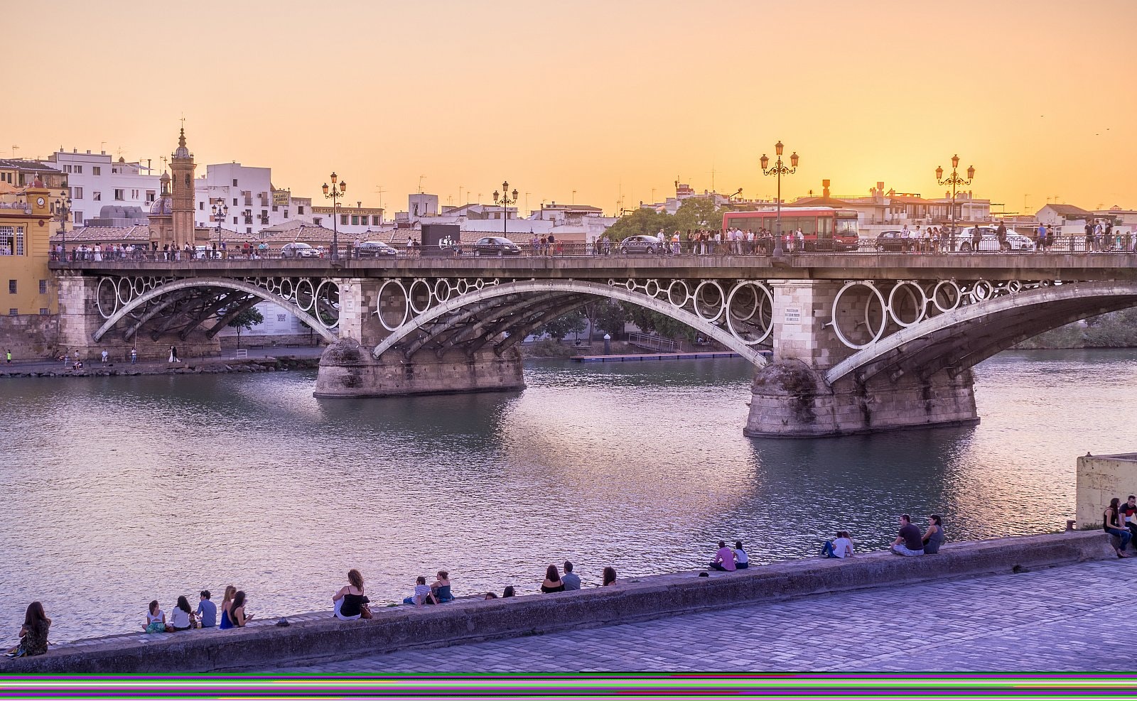 A stone arch bridge across a river at sunset, with buildings beyond and people sitting at the riverside.