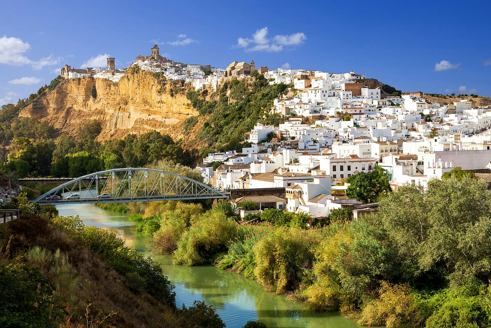Looking out over Arcos de la Frontera, a town of white houses built on a rocky outcrop along the Guadalete river, which is crossed by a tied arch bridge.