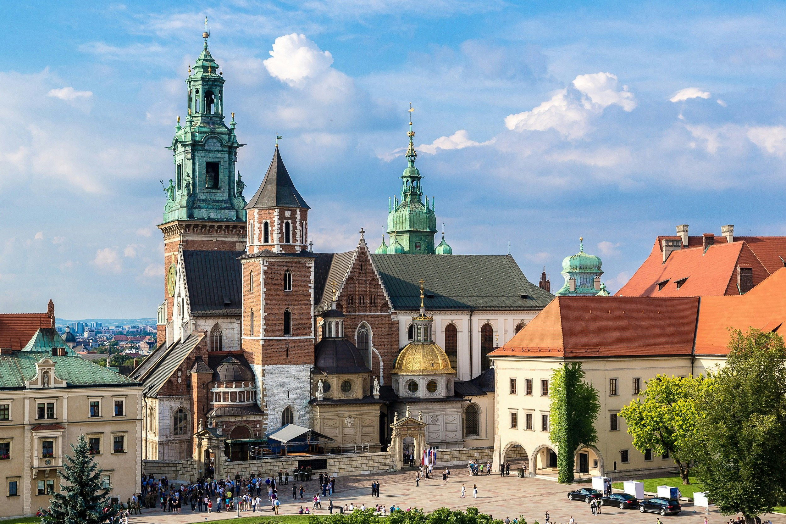 A large cathedral building in Gothic style is surrounded by other grand buildings. People can be seen moving around the grounds at the front