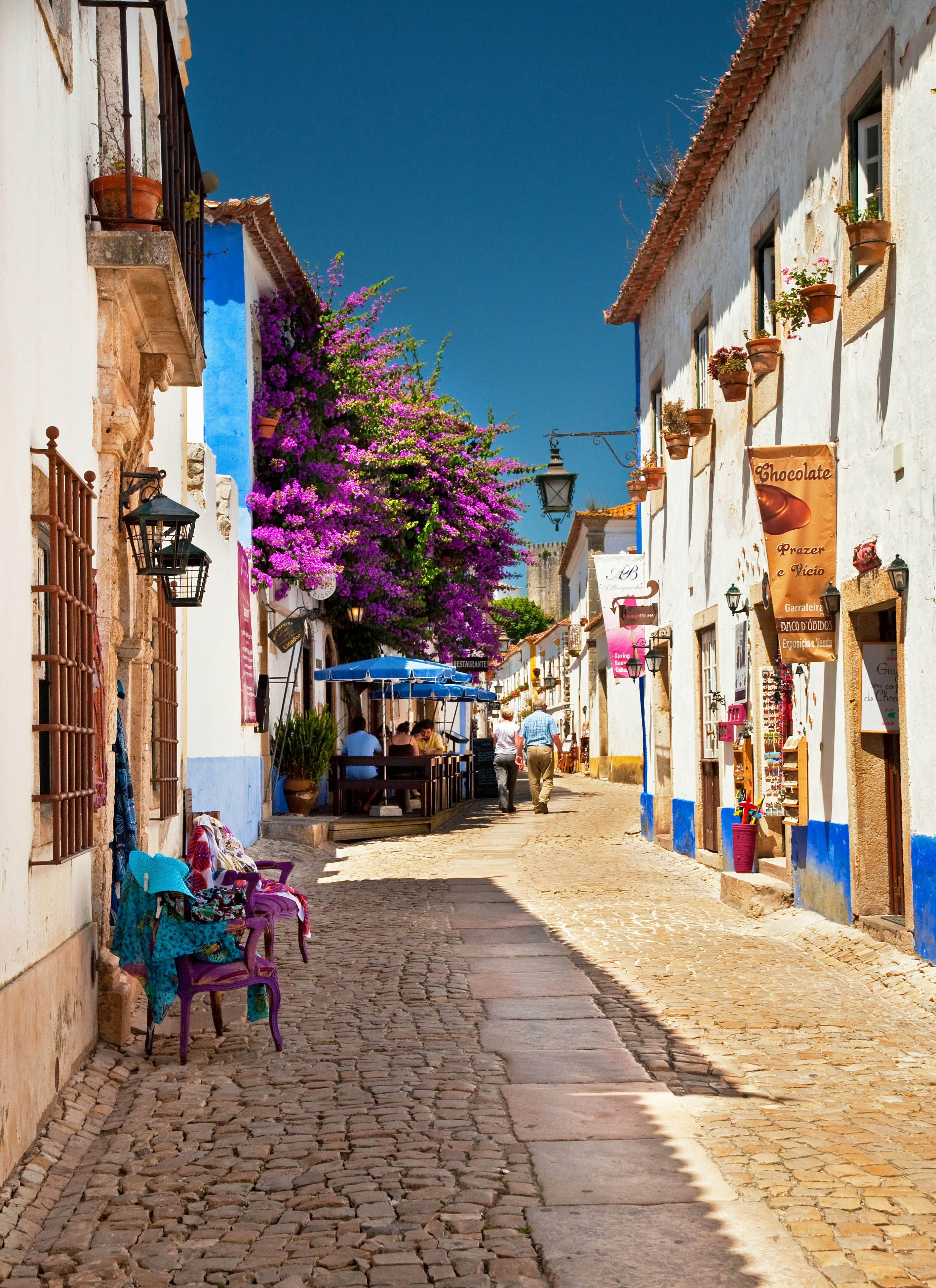 A picturesque, narrow street with sand-coloured cobblestones and whitewashed buildings on either side. Terracotta flowerpots adorn the windows, and bright purple bougainvillea is growing over the azure parasols of a cafe.