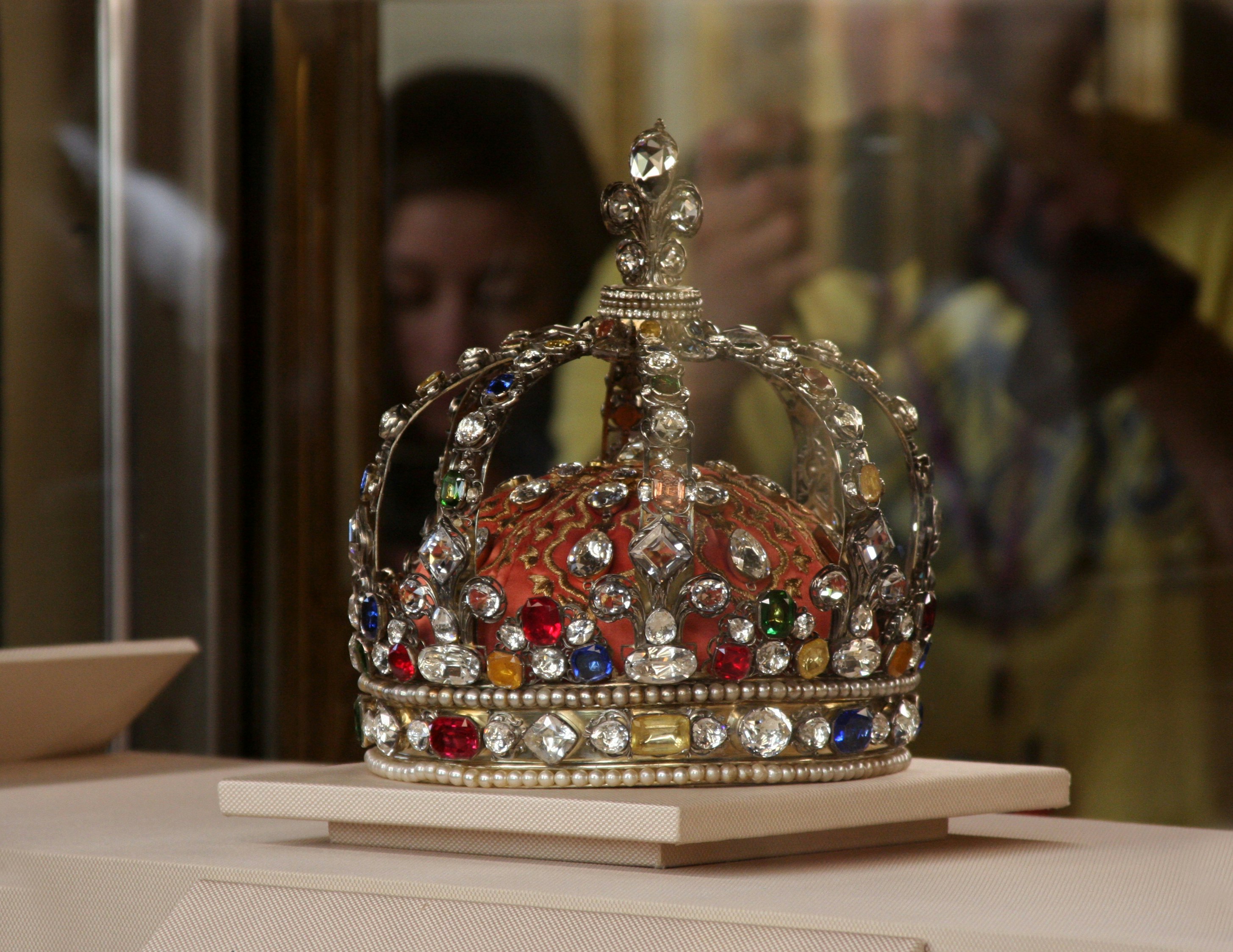 An elaborate crown, encrusted with jewels of different colours, sits on a white display inside a glass case.