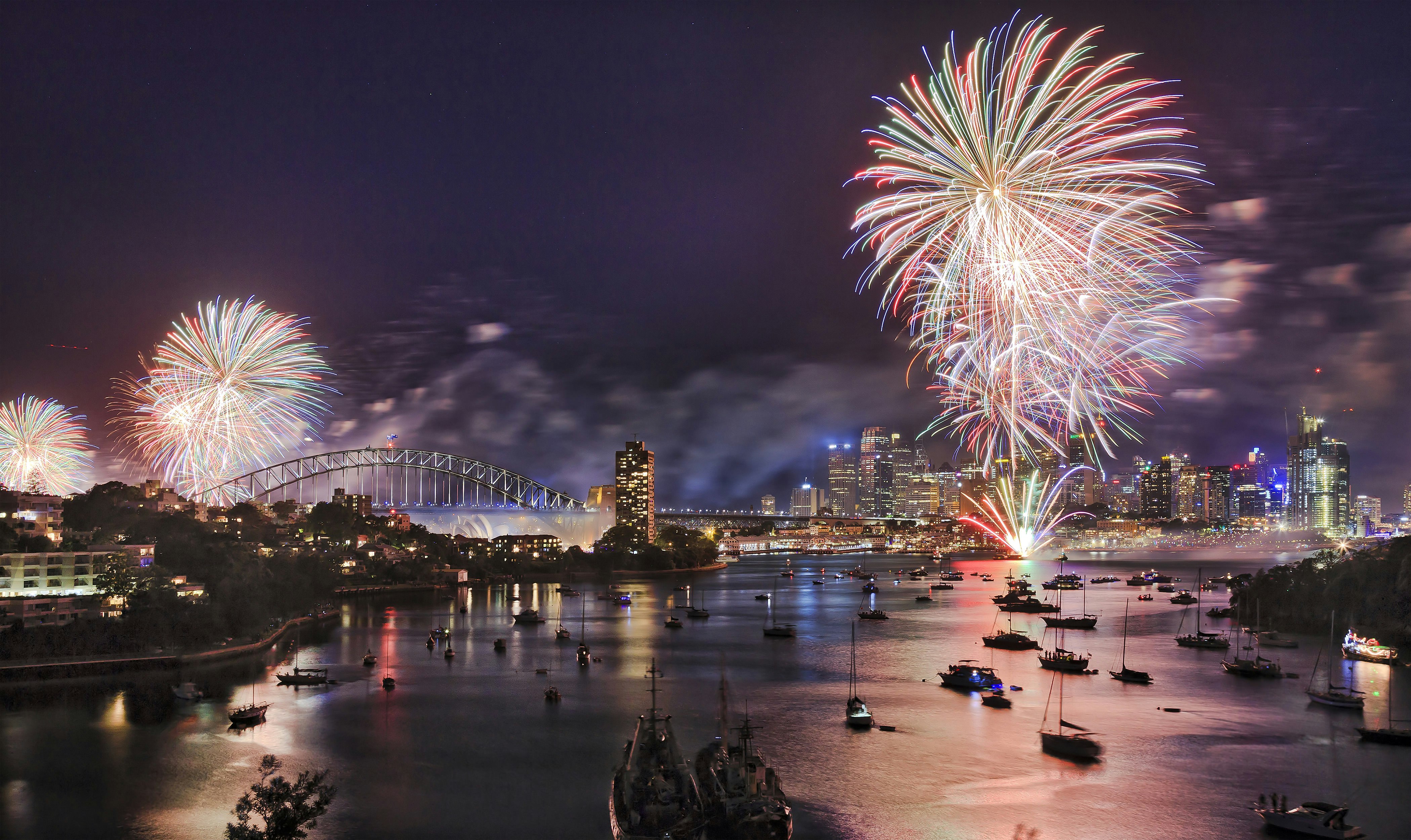 New Year's Eve fireworks explode over Sydney Harbour at night. The harbor is filled with boats, and the Sydney Harbour Bridge and buildings of the CBD can be seen beyond.