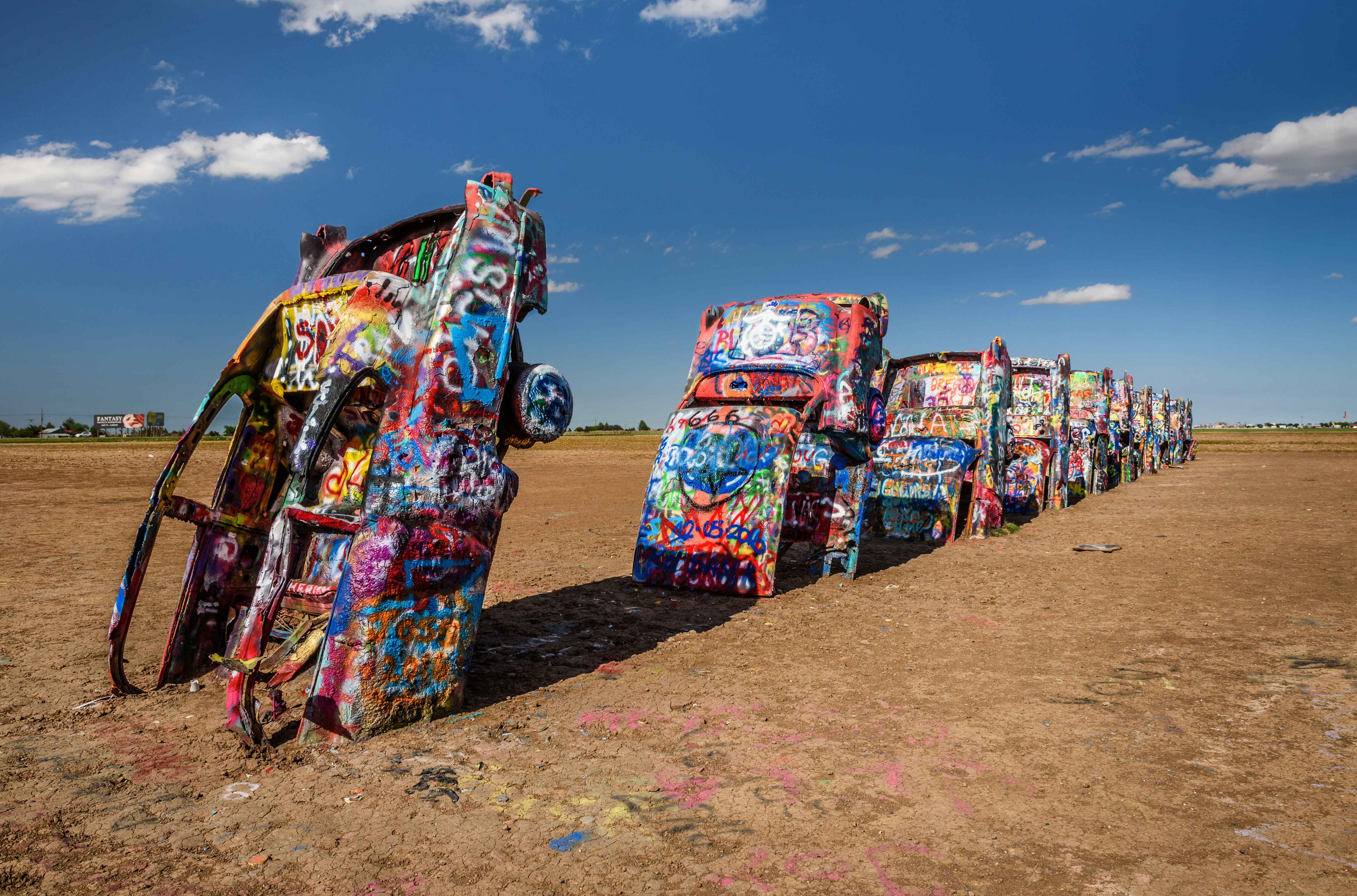 A row of 10 Cadillacs with their hoods buried in the earth stretches out. Each one is covered in colorful marks and graffiti 